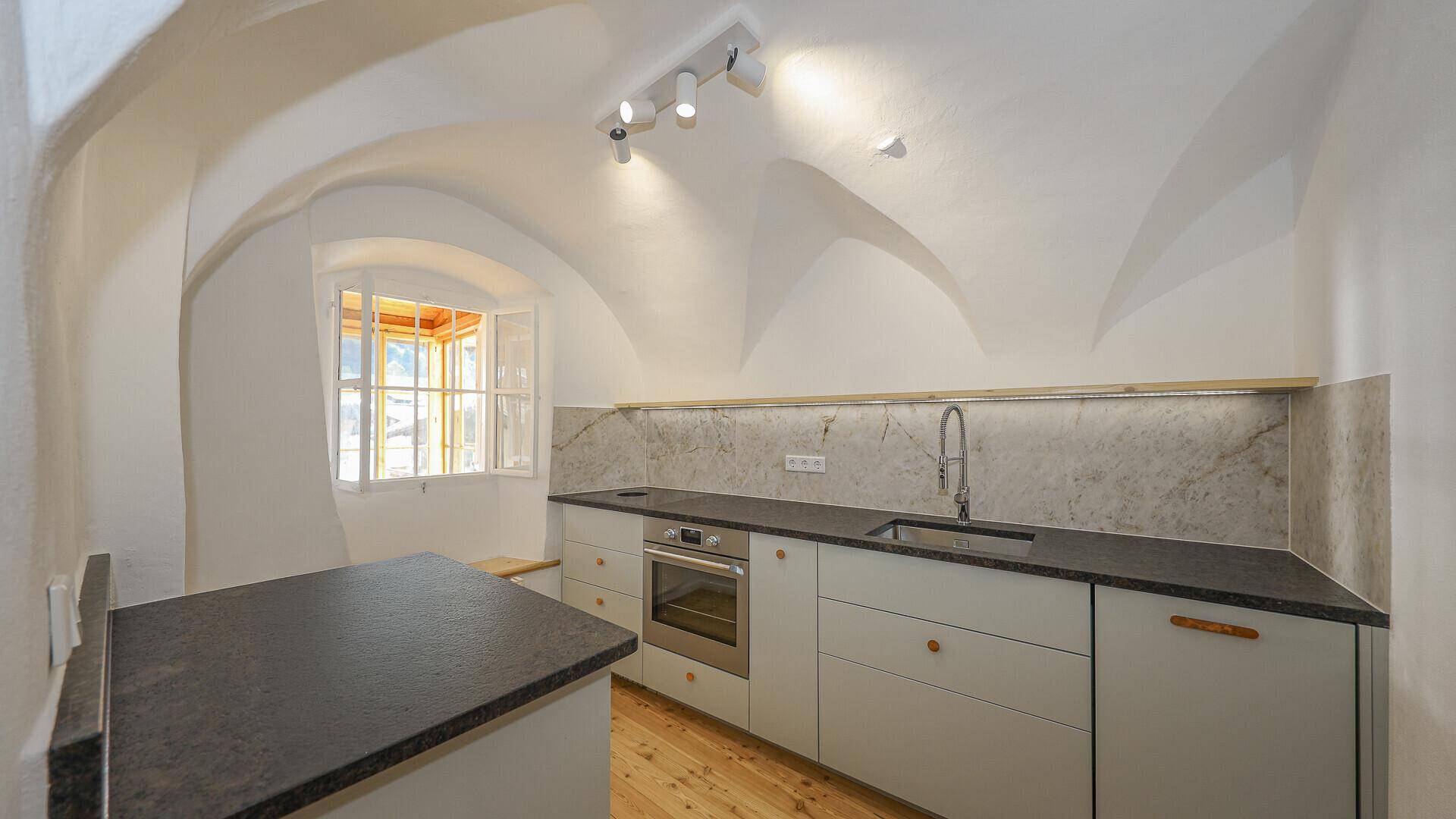Moderne Küche mit Gewölbedecke, hellgrauen Schränken, dunkler Arbeitsplatte und integriertem Backofen.