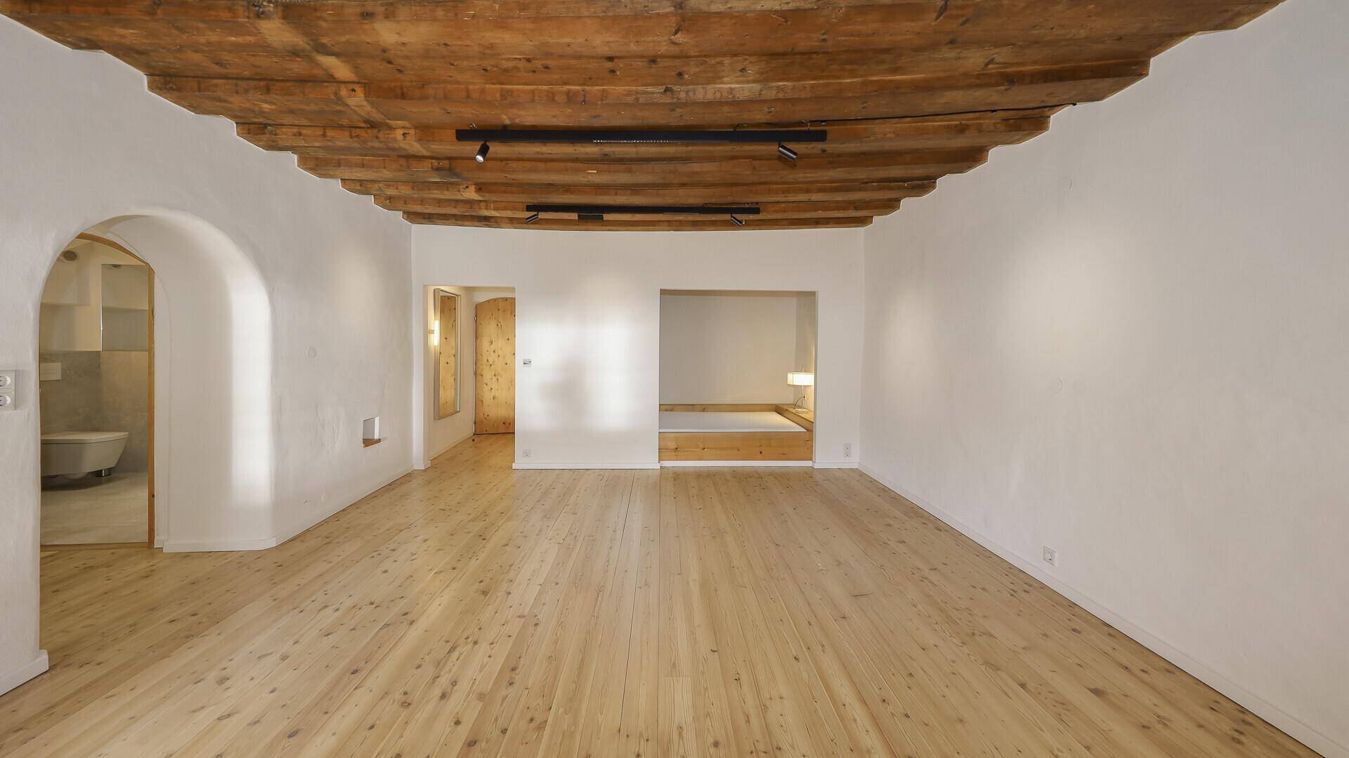 Großzügiger Raum mit beeindruckender Holzbalkendecke, hellem Dielenboden und Zugang zu einem Badezimmer.
