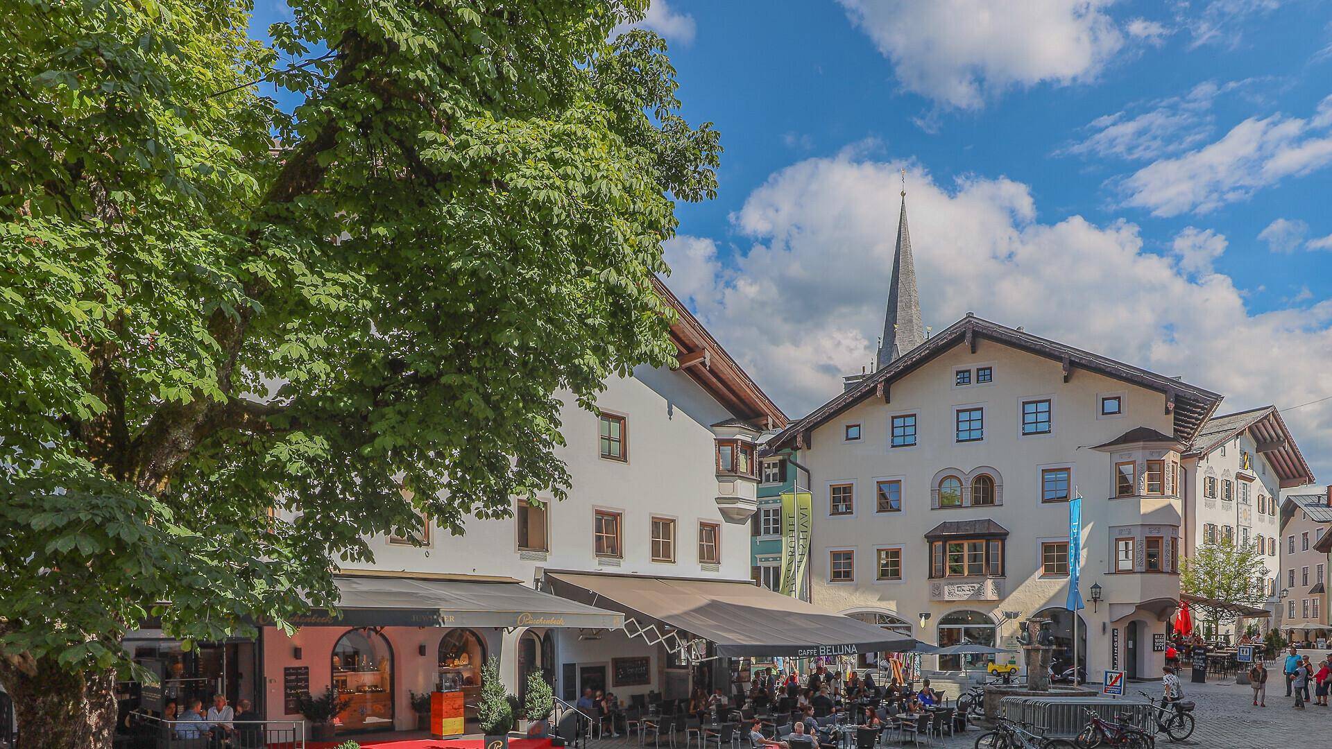 Lebhafter Stadtplatz mit traditionellen Gebäuden, Außengastronomie und einem Kirchturm im Hintergrund.
