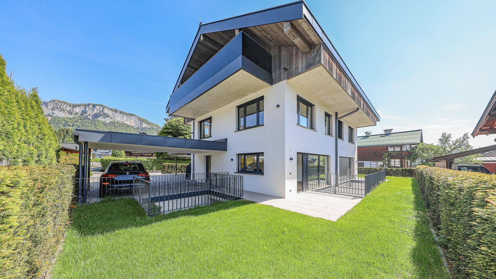 Modernes Einfamilienhaus mit Carport, Garten und Bergblick unter blauem Himmel.