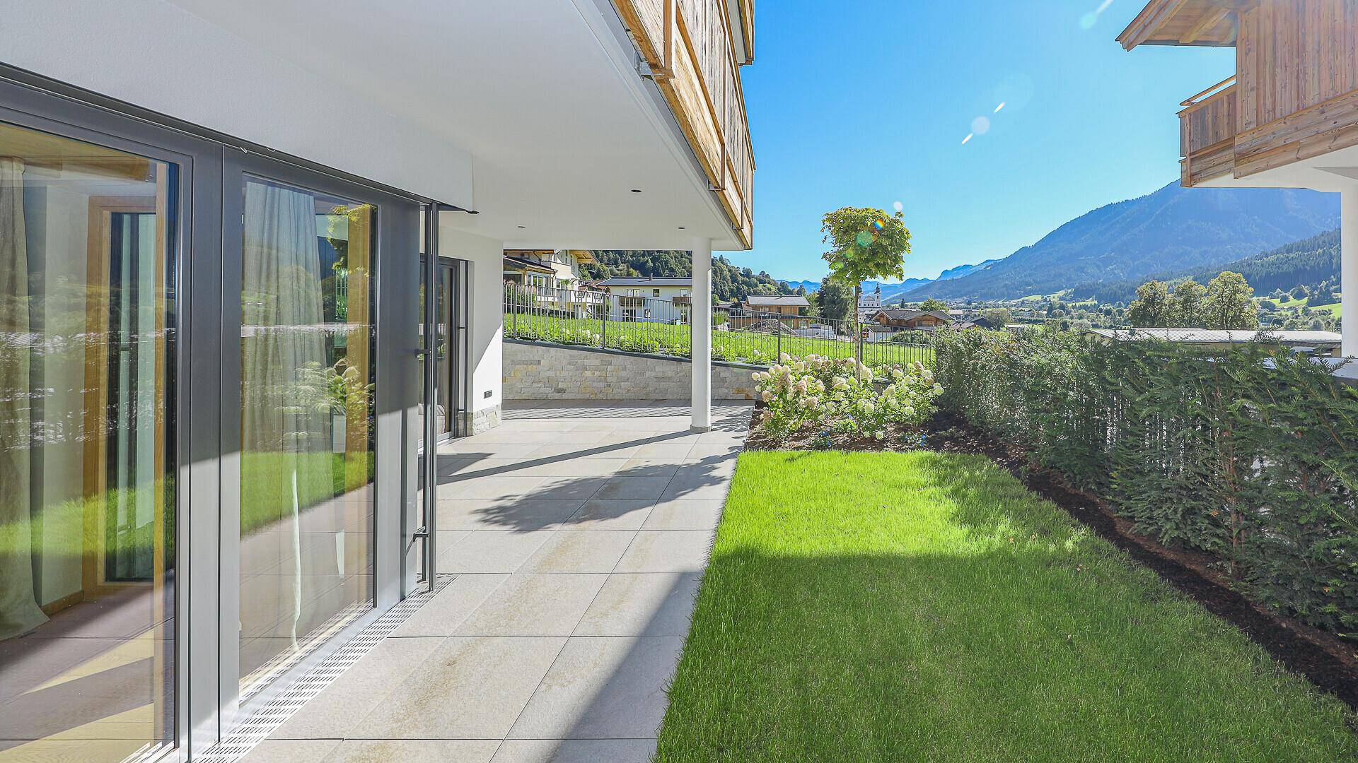 Sonniger Gartenbereich mit großer Terrasse, Rasen und Blick auf die umliegende Berglandschaft.