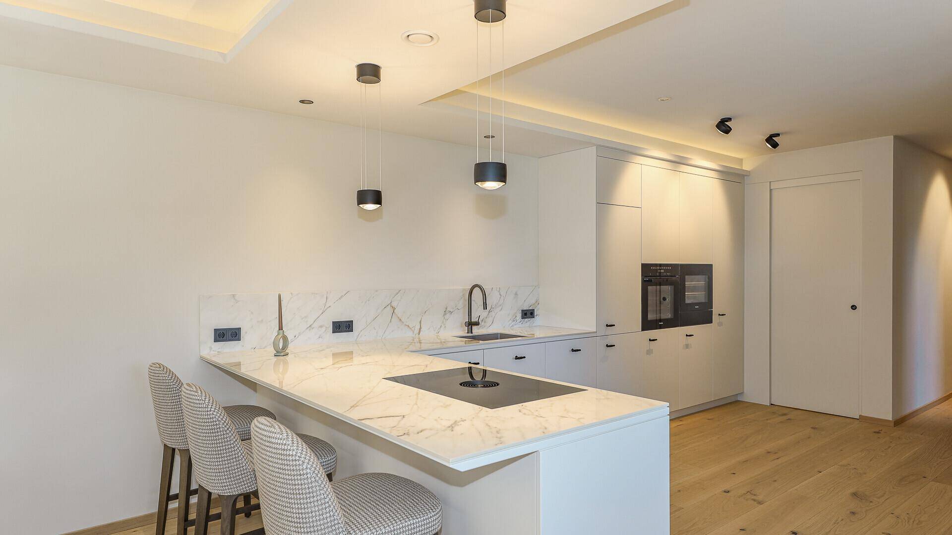 Elegante, moderne Küche mit weißer Arbeitsplatte, integrierten Geräten und Barhockern für schnelles Essen.