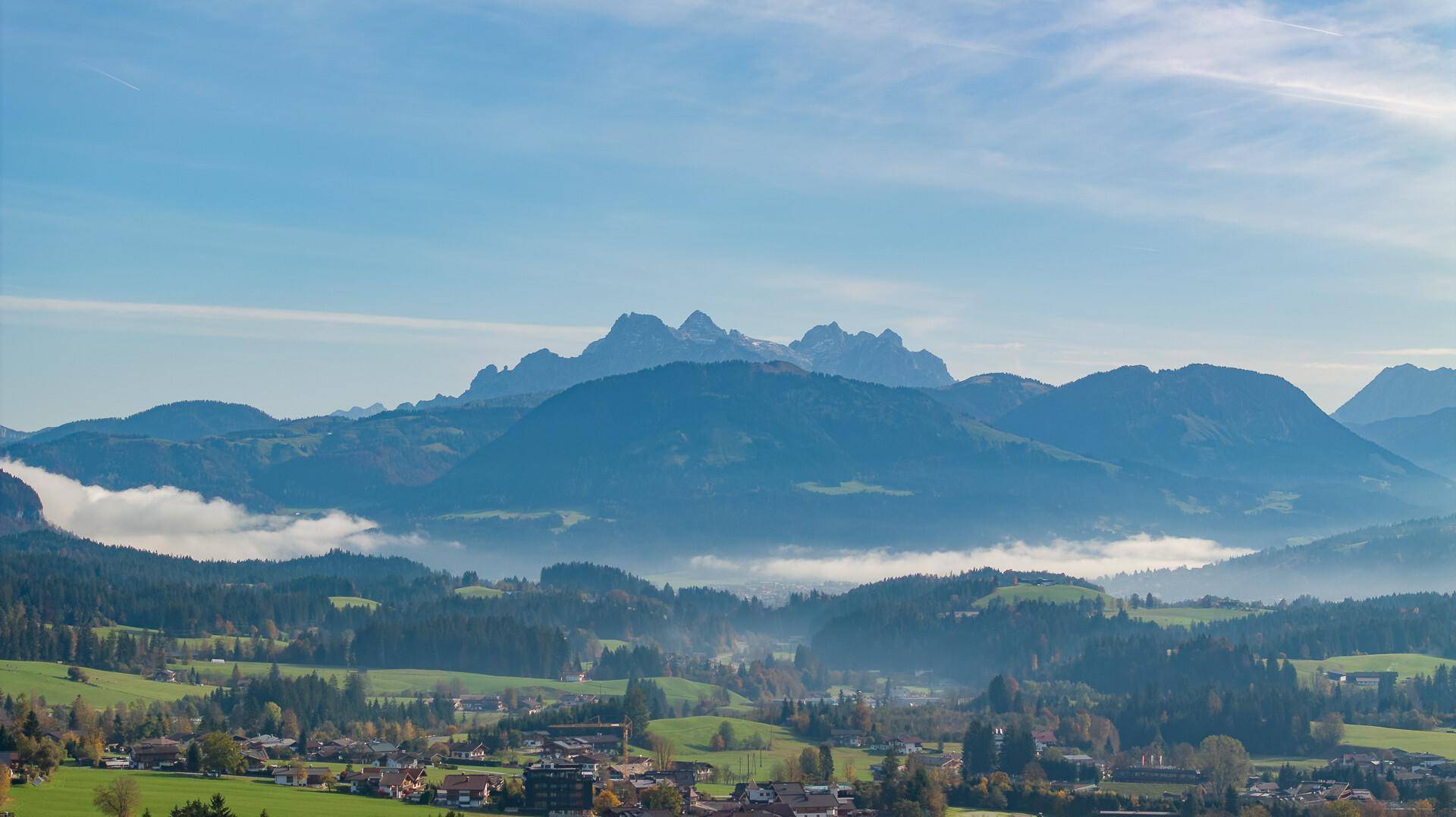 Weitläufige Landschaft mit sanften Hügeln, Wäldern und einem Dorf im Tal, umgeben von Bergen.