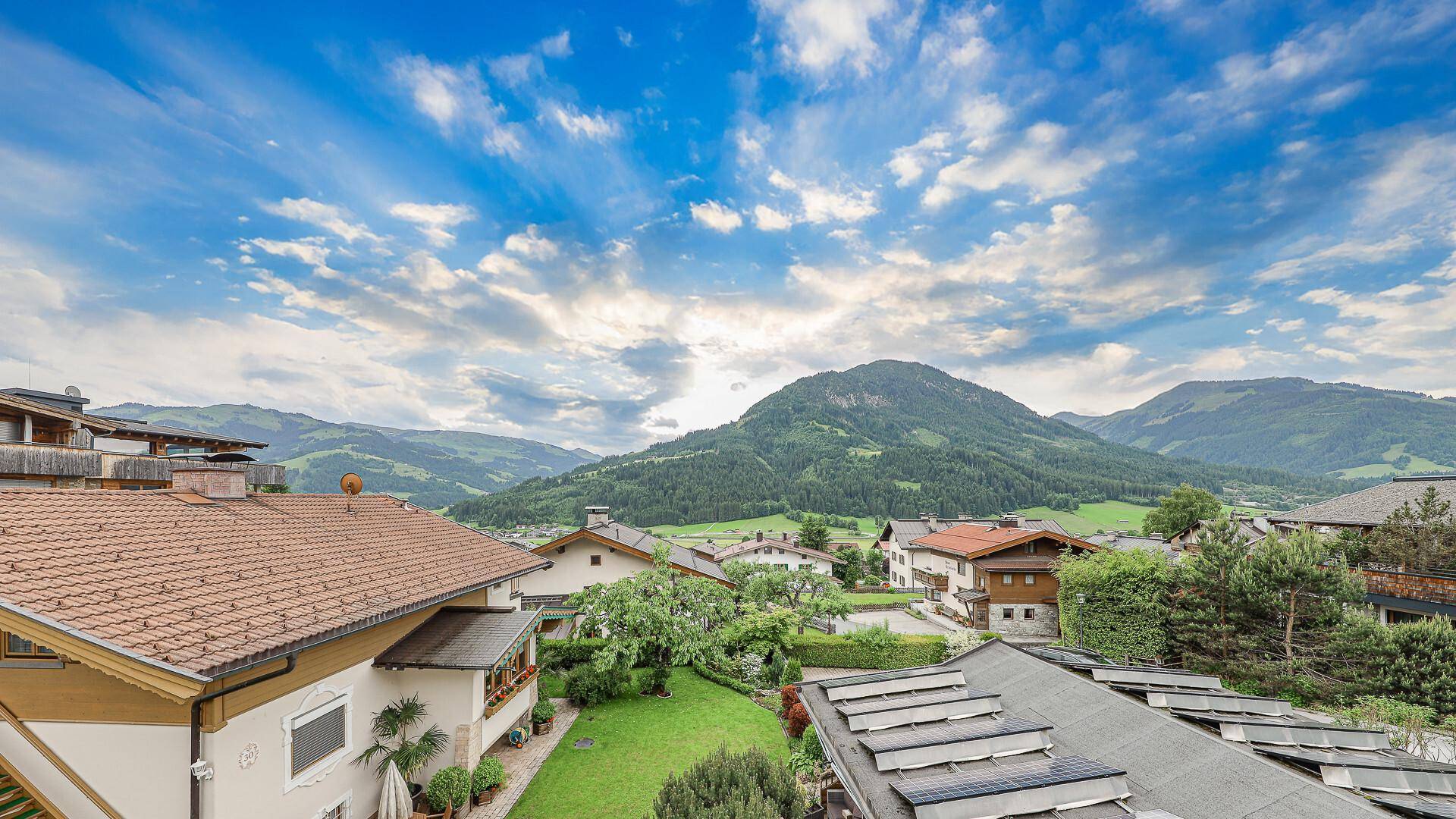 Panoramablick über Dächer mit Solaranlagen und einer weiten grünen Berglandschaft unter blauem Himmel.