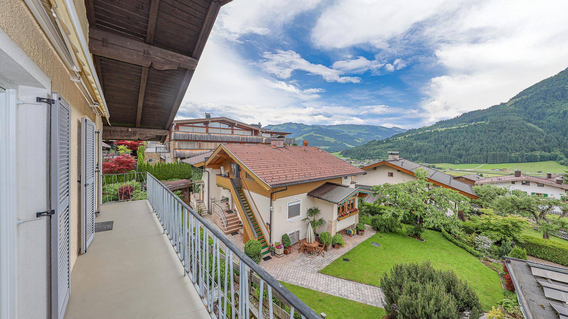 Blick vom Balkon auf eine Terrasse mit Sitzgelegenheiten und einen gepflegten Garten mit Bergblick.