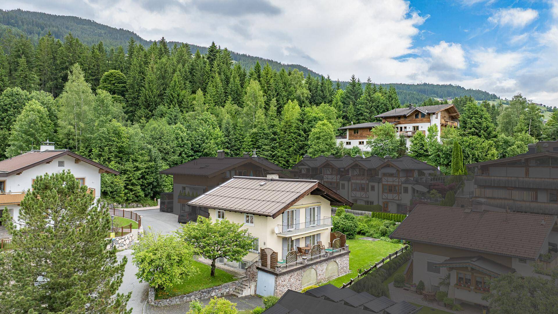 Luftaufnahme eines Hauses mit heller Fassade und braunem Dach, eingebettet in eine grüne Berglandschaft.