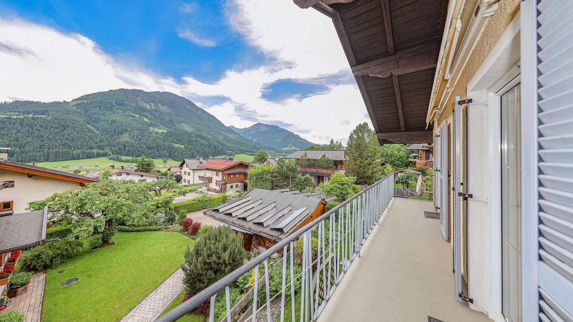Blick vom Balkon auf die grüne Landschaft mit Häusern und Bergen im Hintergrund unter blauem Himmel.