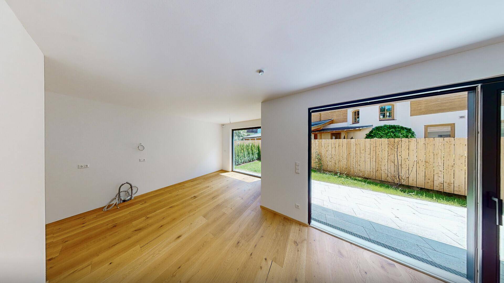 Geräumiger, heller Raum mit Holzboden und Zugang zu einer gepflasterten Terrasse und dem Garten.