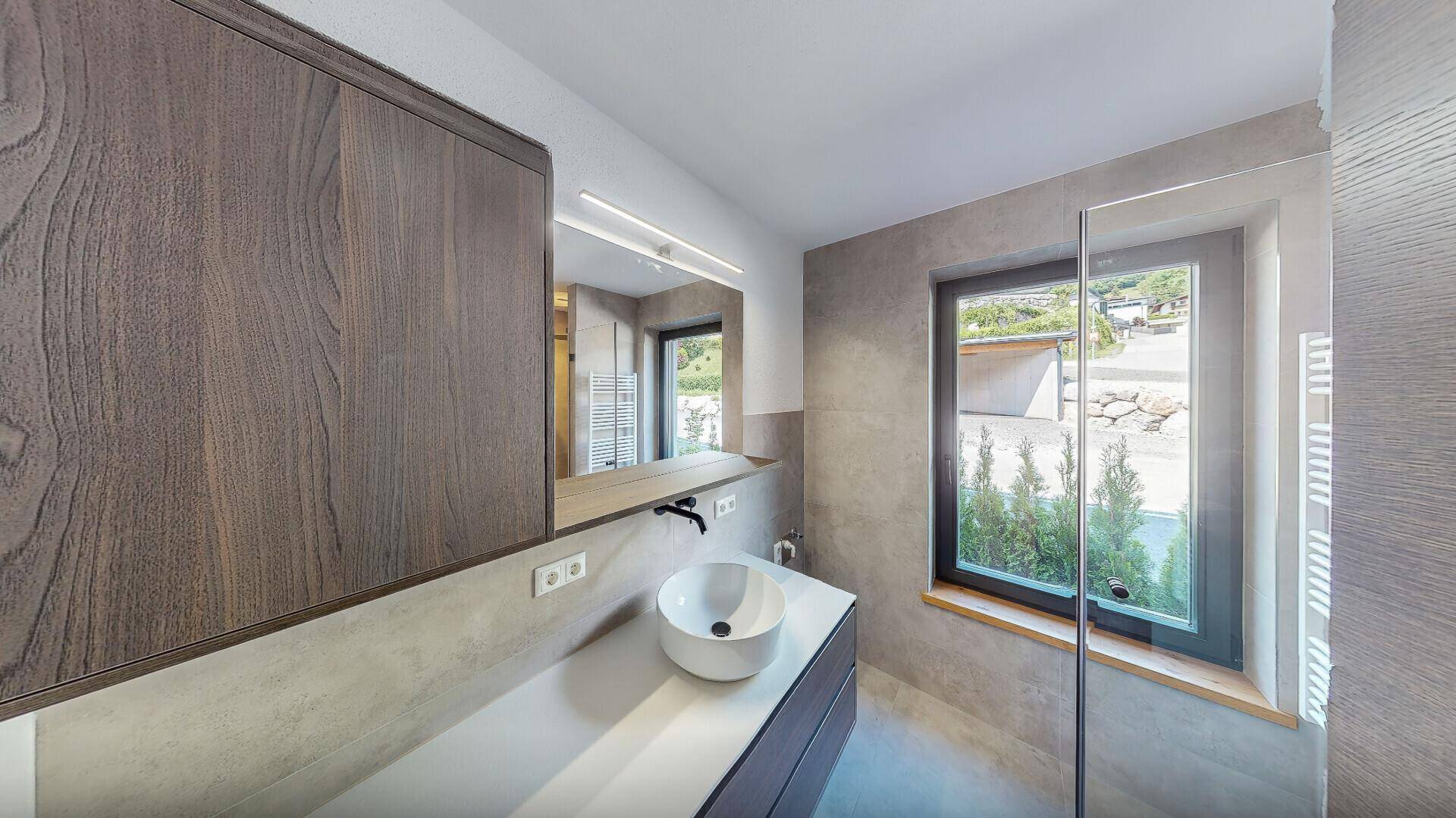 Elegantes Badezimmer mit grauen Fliesen, Einzelwaschbecken, großem Spiegelschrank und Fenster.