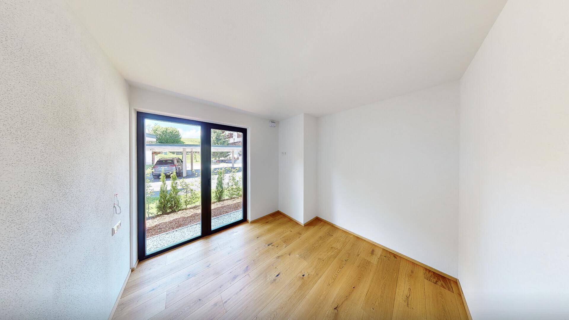 Heller Raum mit Holzboden und großen Glastüren, die einen Blick auf den Außenbereich und Carport ermöglichen.