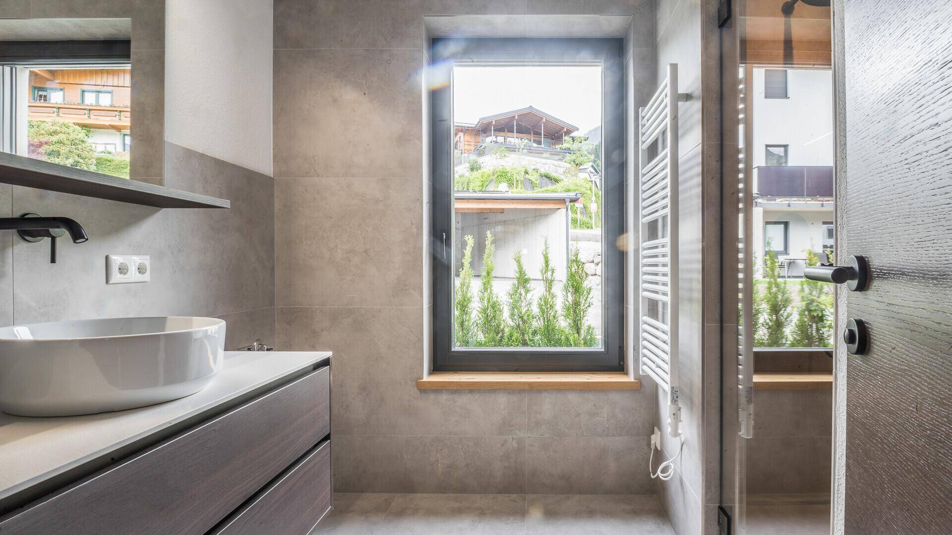 Modernes Badezimmer mit grauen Fliesen, Einzelwaschbecken, Duschbereich und einem Fenster für Tageslicht.