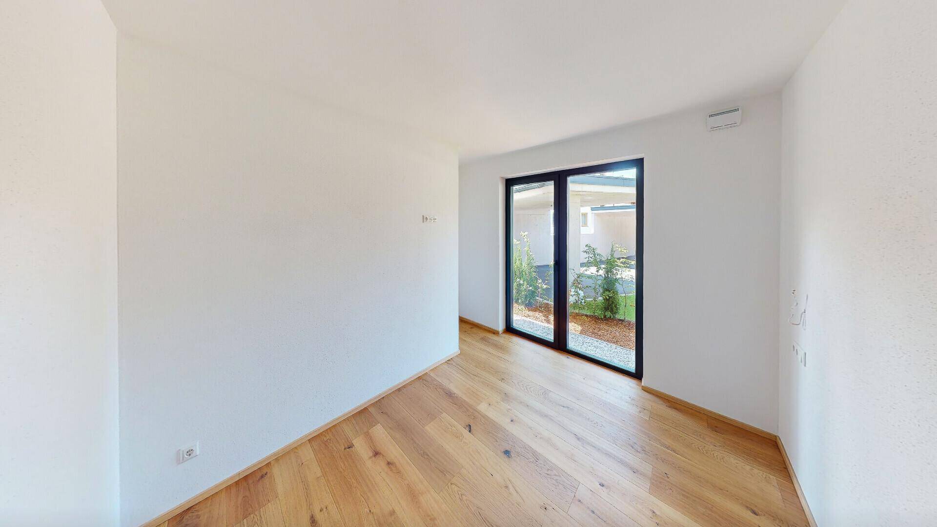 Heller, unmöblierter Raum mit Holzboden und großen Glastüren, die Zugang zum Außenbereich bieten.