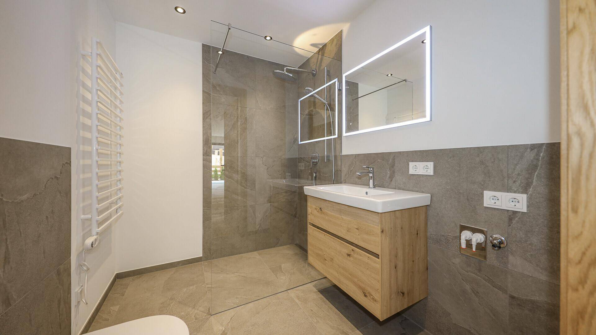 Modernes Badezimmer mit begehbarer Dusche, Holzwaschtisch und beleuchteten Spiegeln.