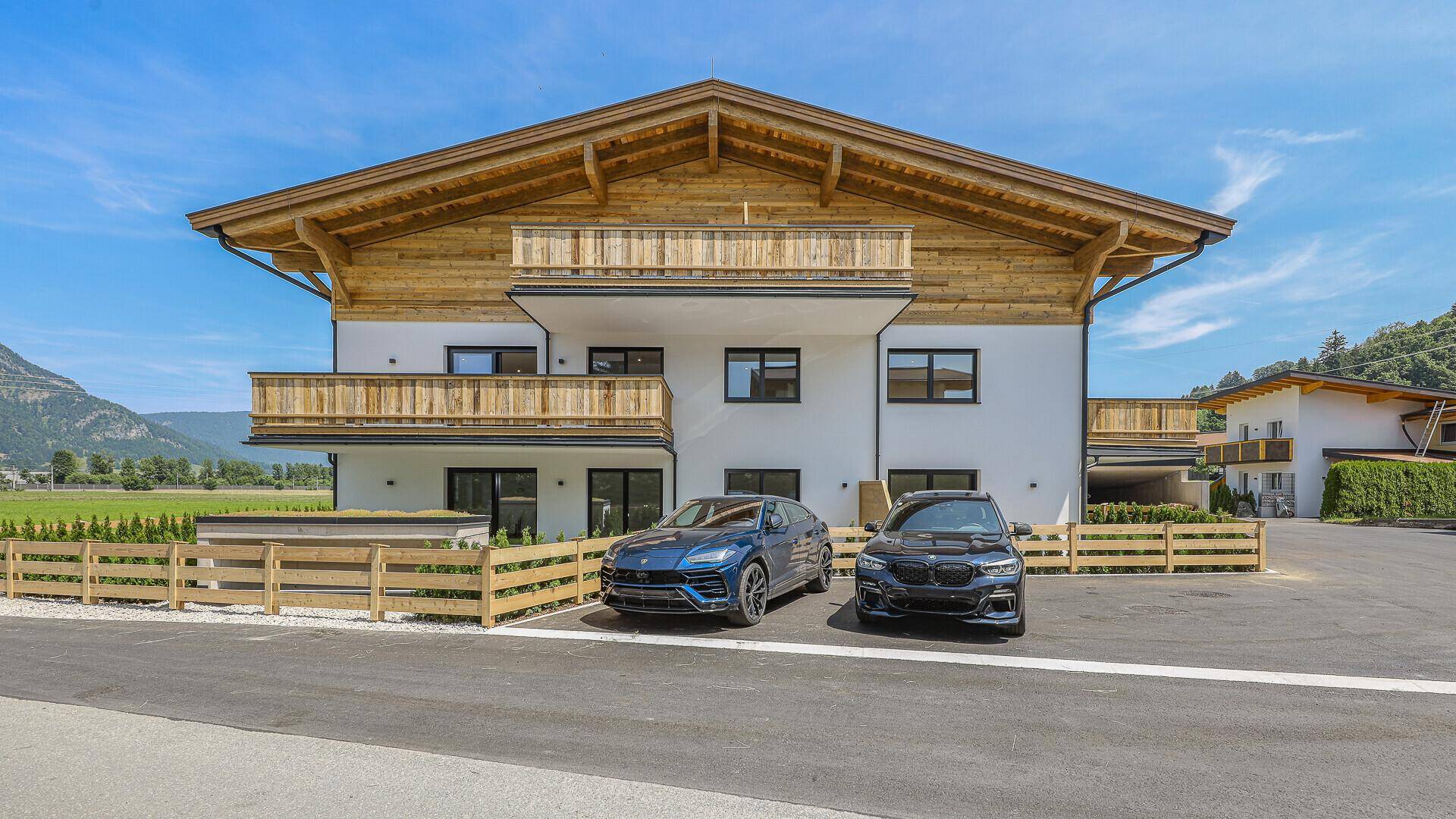 Frontansicht eines modernen Neubaus im alpinen Stil mit geparkten Autos und Holzzaun.
