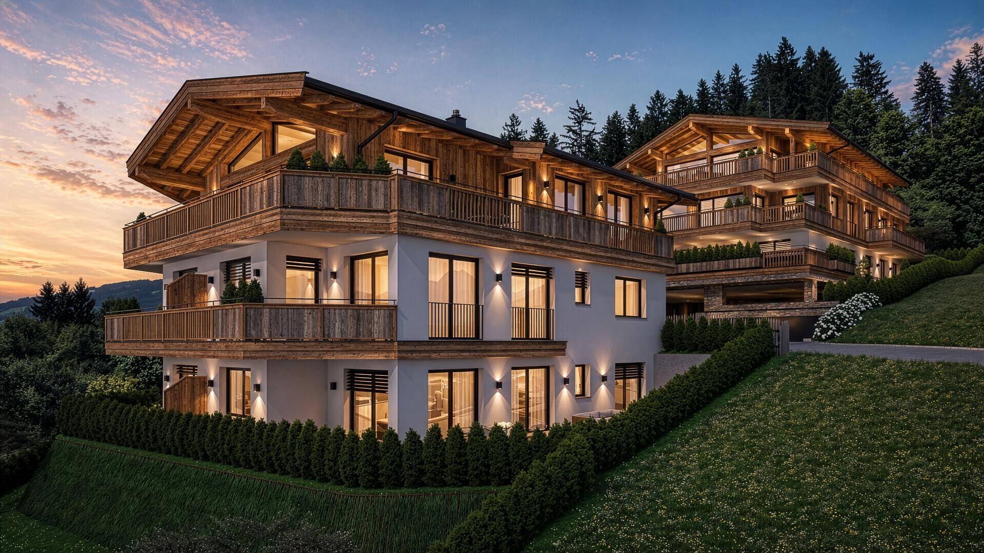 Zwei moderne Chalets in Hanglage bei Sonnenuntergang, mit beleuchteten Fassaden und Balkonen, umgeben von Natur.
