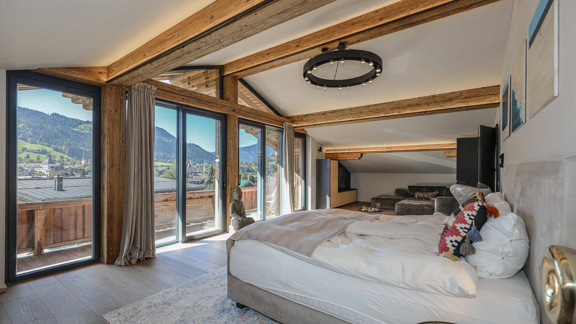 Helles Schlafzimmer mit Holzbalken, großen Fenstern und Panoramablick auf Berge und Stadt.