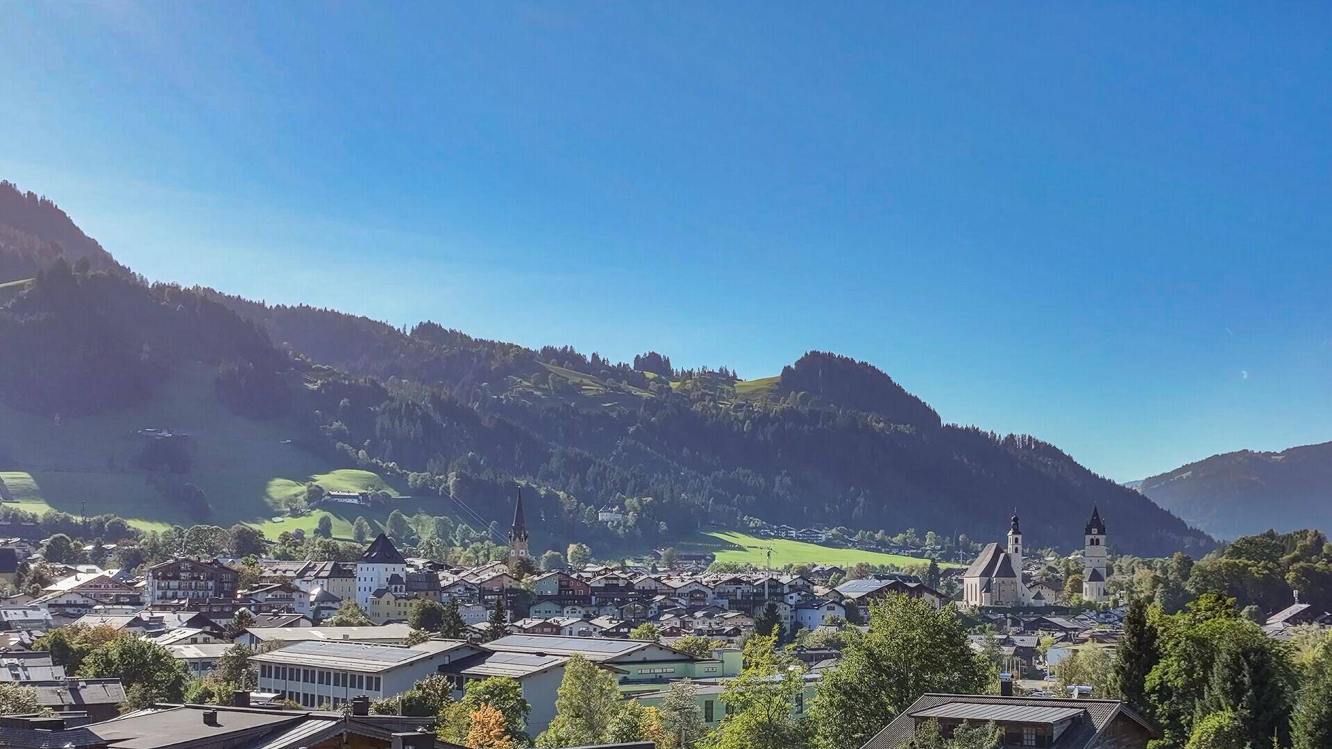 Panoramablick auf eine malerische Stadt im Tal, umgeben von grünen Bergen unter blauem Himmel.