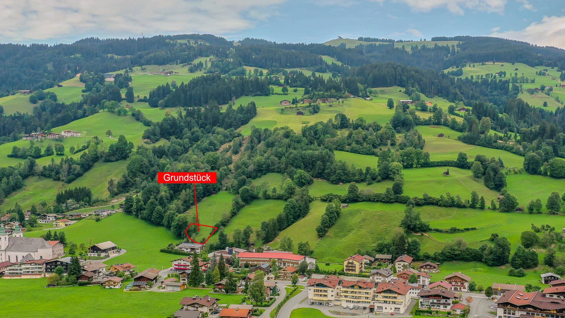 Luftaufnahme eines Grundstücks am Hang, eingebettet in die grüne Hügellandschaft eines Dorfes.