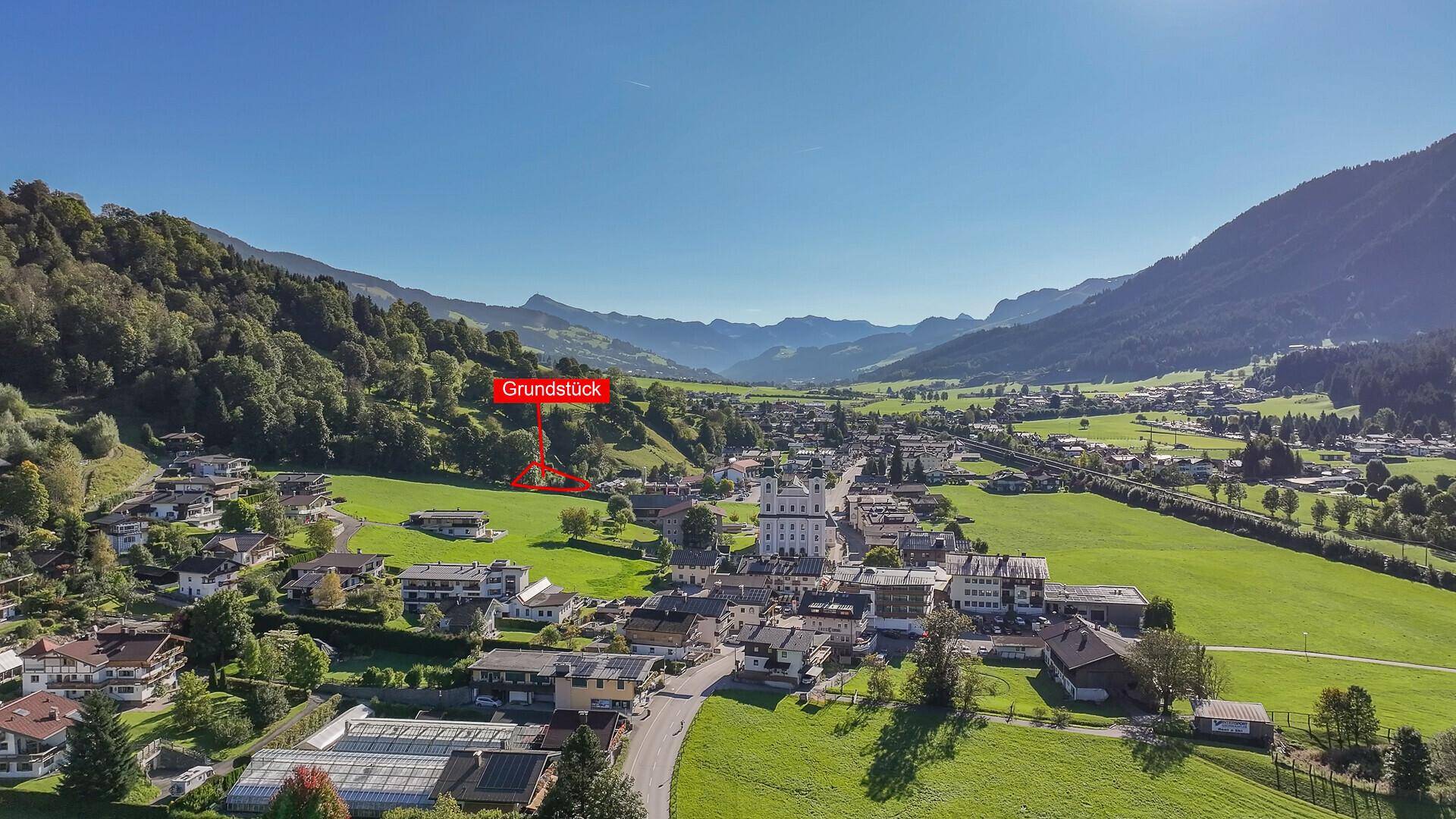 Luftbild eines Baugrundstücks mit Panoramablick auf das Dorf und die umliegende Bergwelt.