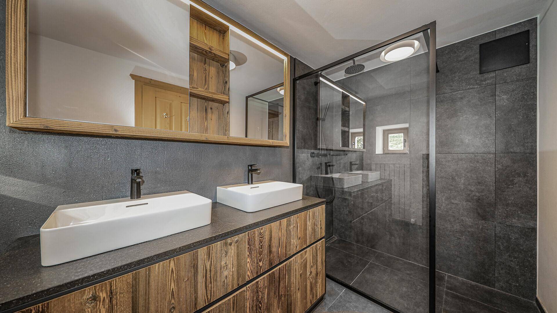 Modernes Badezimmer mit Doppelwaschbecken, großer Dusche und dunklen Fliesen.