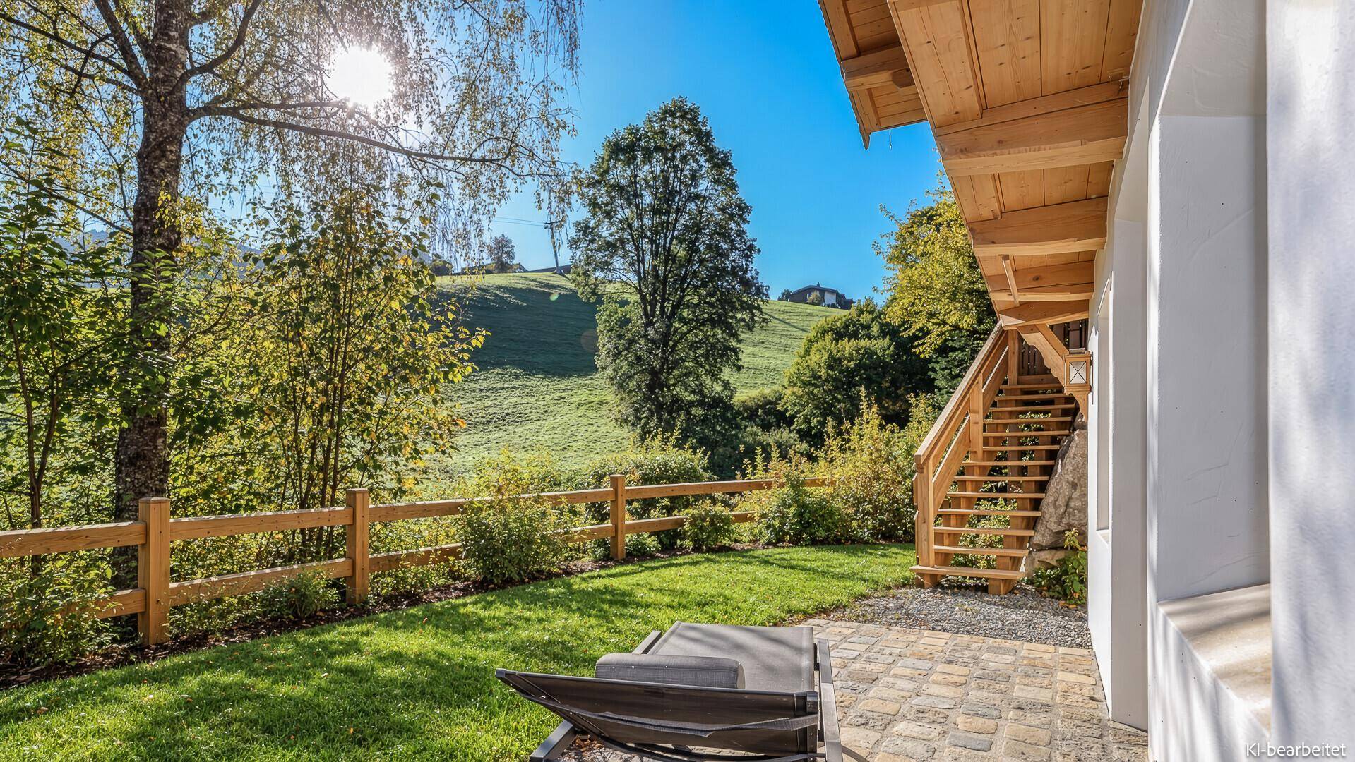 Sonnige Rasenfläche mit gepflastertem Bereich und Blick auf die grüne Hügellandschaft.