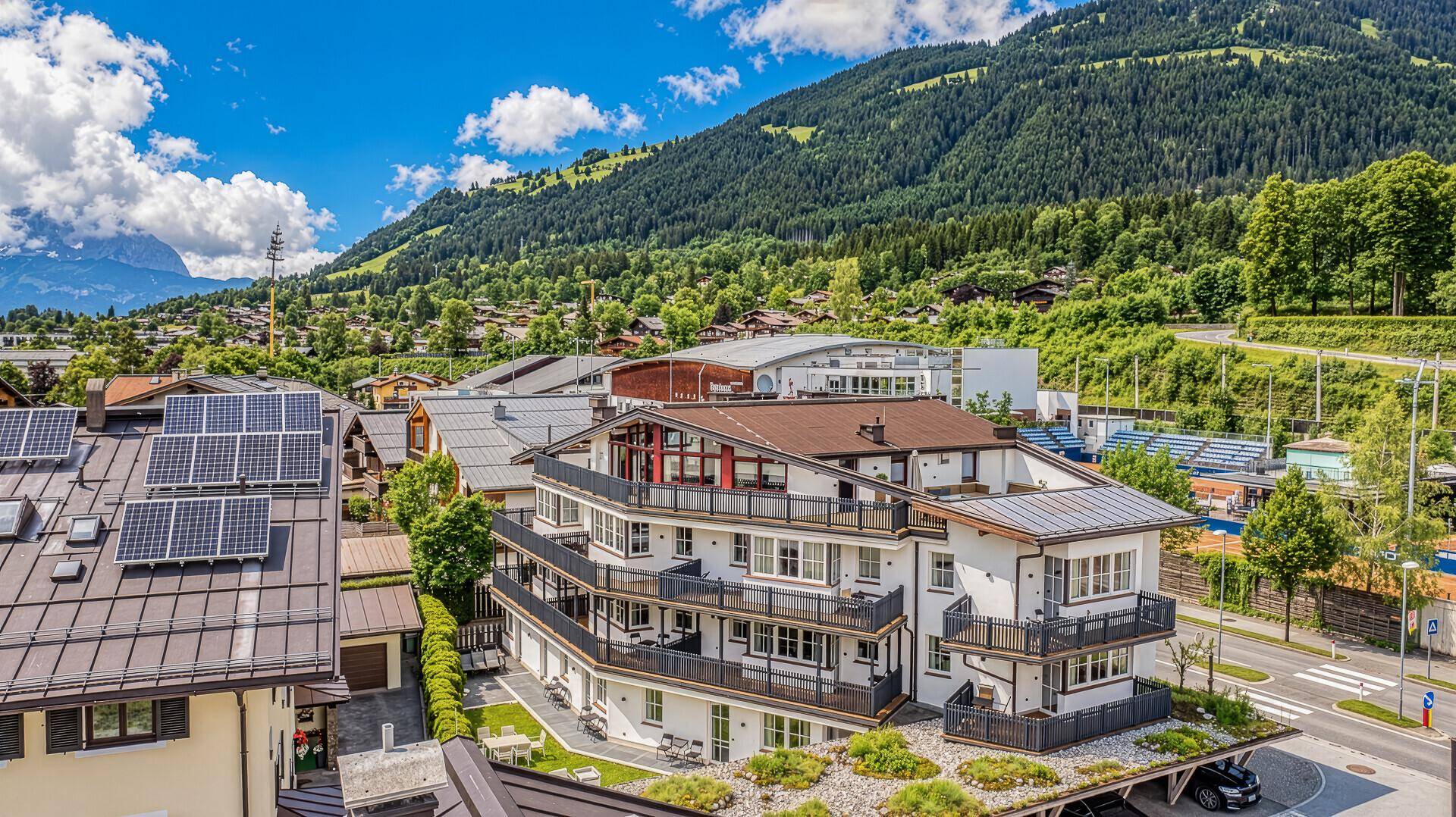 Luftaufnahme der Wohnanlage im Sommer mit grünen Bergen und Photovoltaikanlage auf dem Dach.