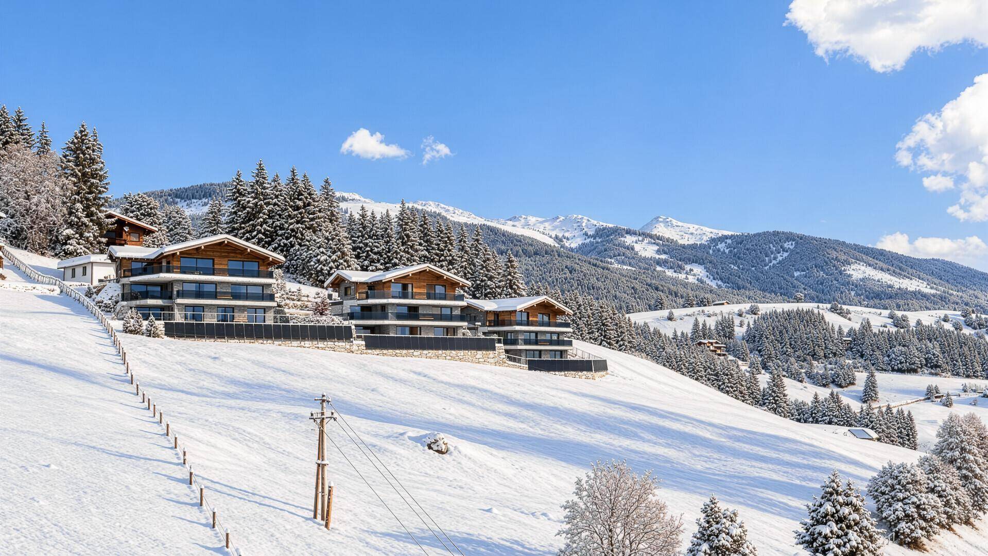 Mehrere moderne Chalets in einer weiten, verschneiten Berglandschaft unter blauem Himmel.