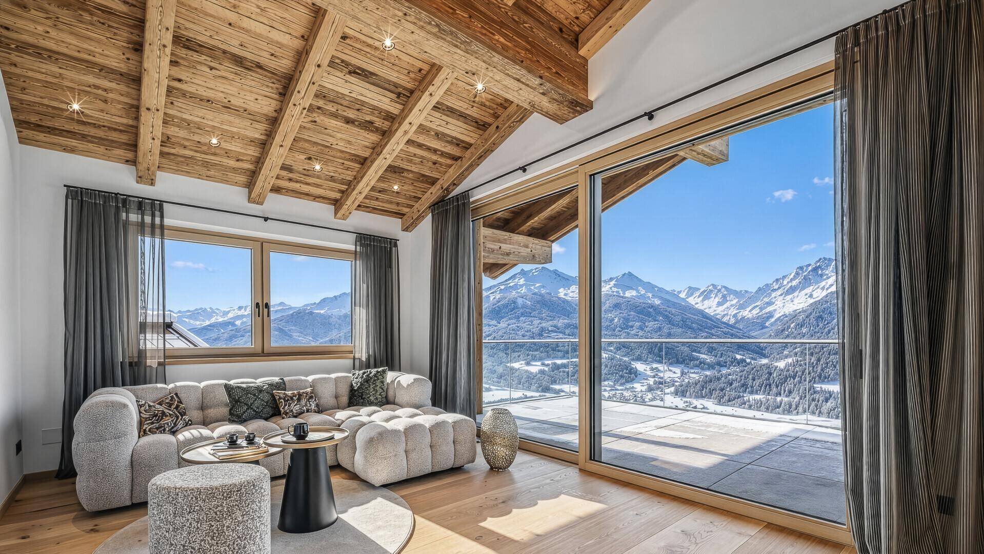 Gemütliches Wohnzimmer mit bequemer Couch, Holzbalkendecke und weitem Blick auf die verschneiten Berge.