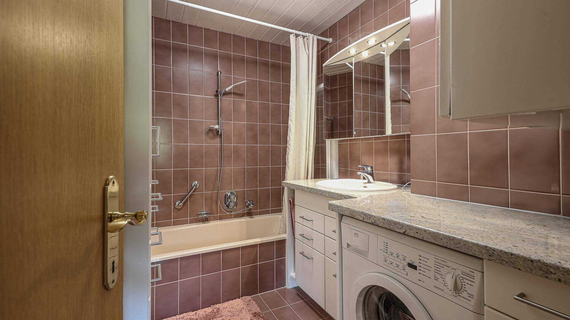 Funktionales Badezimmer mit braunen Fliesen, Badewanne mit Duschfunktion und Waschmaschine.