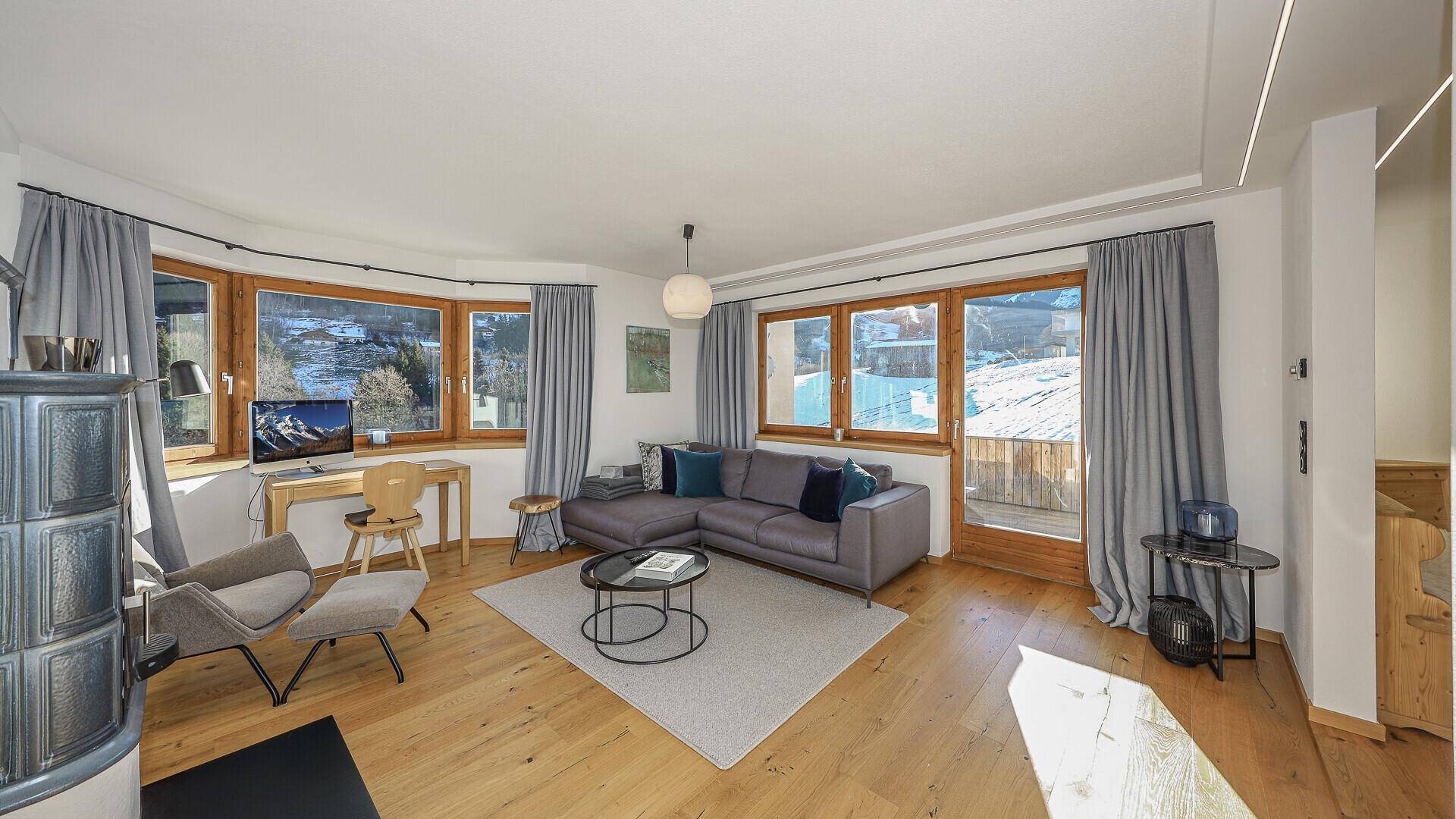 Helles Wohnzimmer mit Holzboden, Ecksofa und einem Arbeitsbereich am Erkerfenster mit Bergblick.