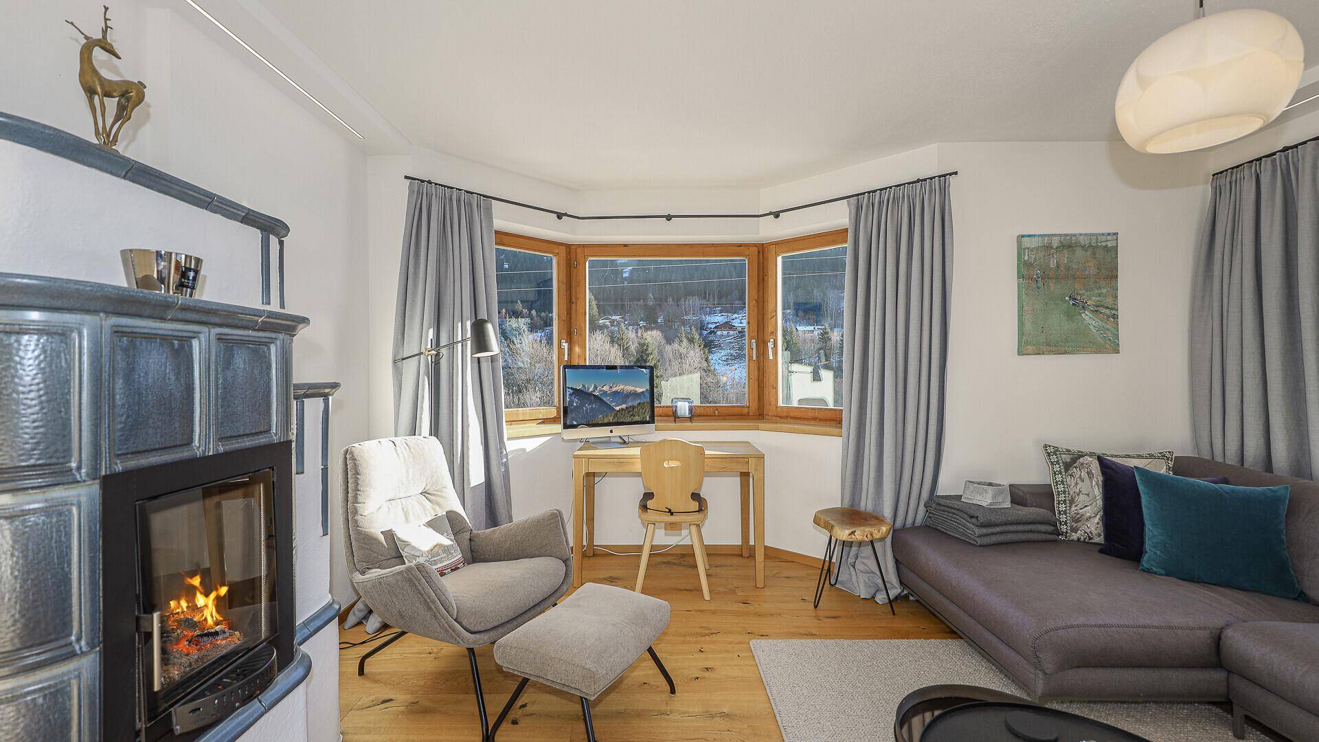 Detailansicht des Wohnzimmers mit Kachelofen, Schreibtisch am Erkerfenster und Blick auf die verschneite Umgebung.