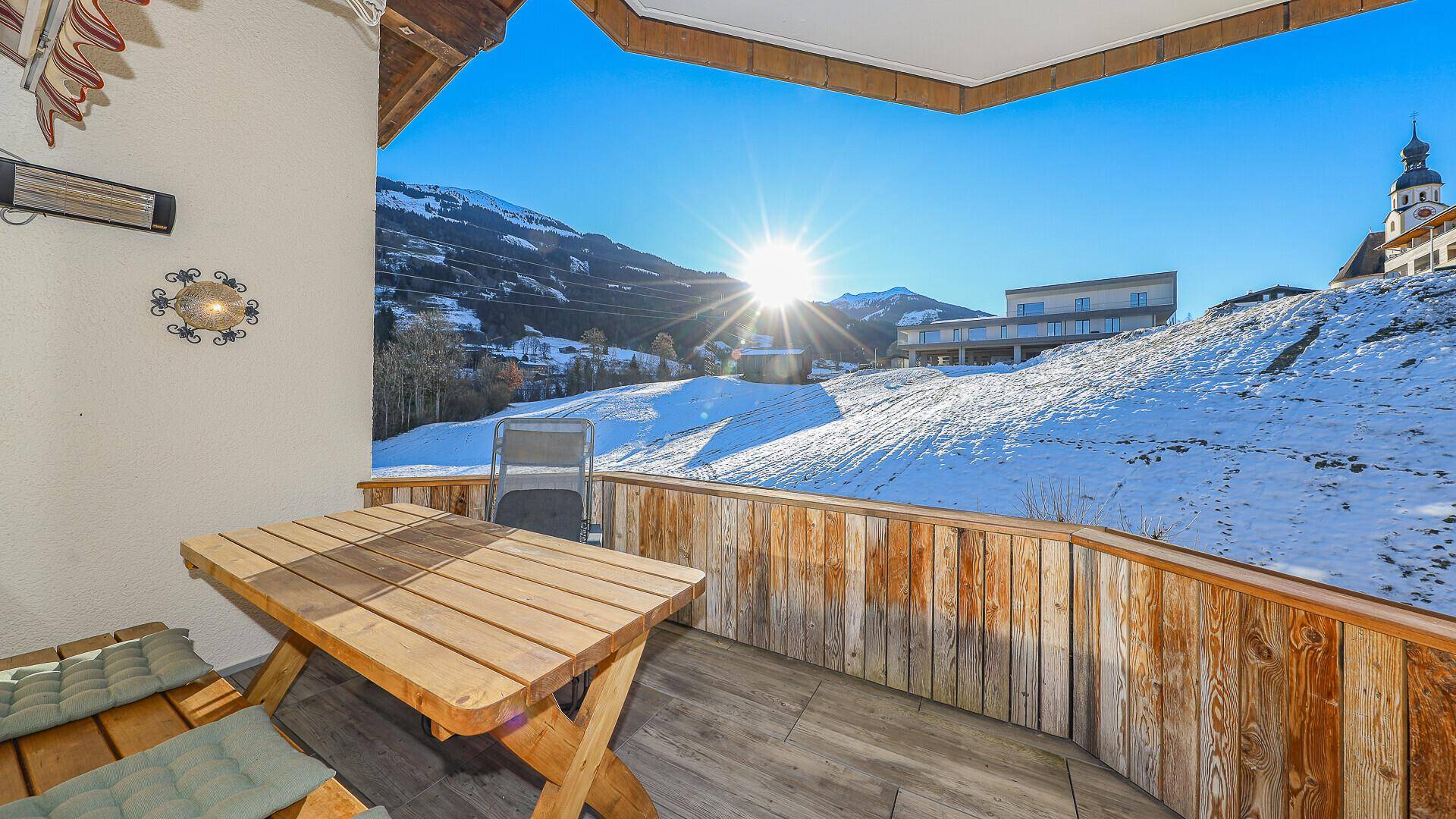 Sonniger Balkon mit Holztisch und Stühlen, bietet einen weiten Blick auf die verschneiten Berge.