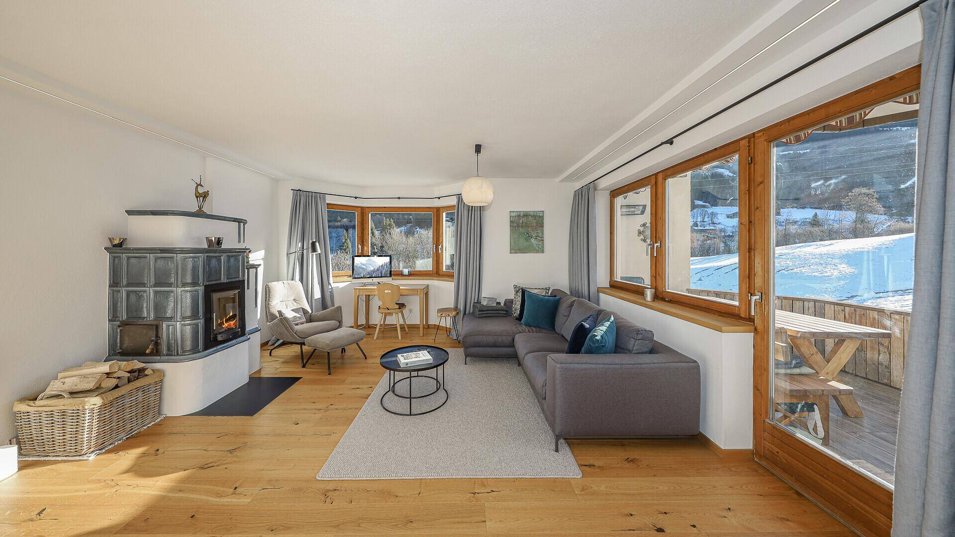 Gemütliches Wohnzimmer mit Kachelofen, Holzboden und großzügigen Fenstern mit Blick auf die verschneite Landschaft.