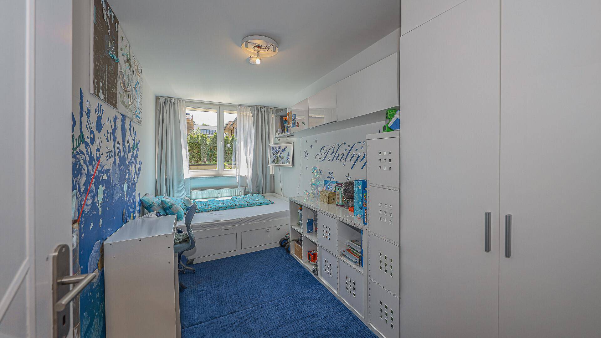 Kinderzimmer mit blauem Teppich, eingebautem Bett und Stauraum.
