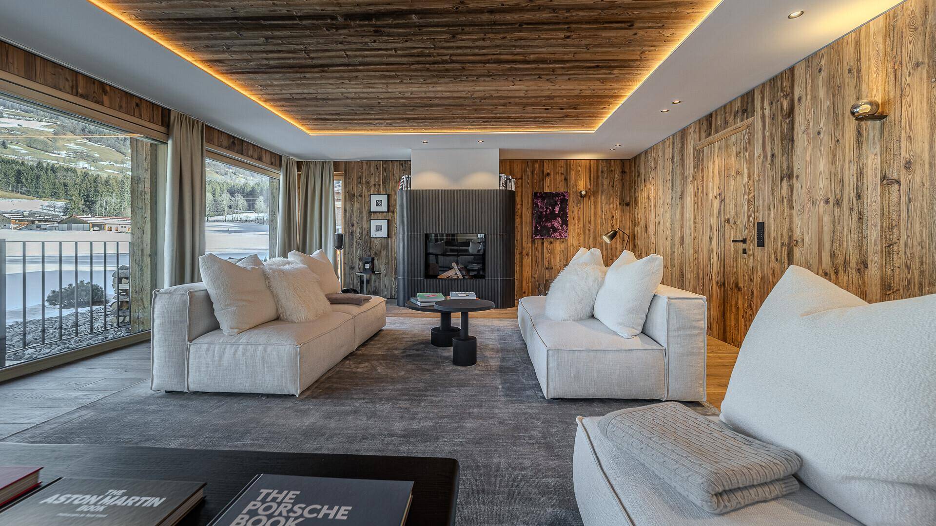 Stilvoller Wohnraum mit Kamin, Holzwänden und großen Fenstern mit Blick auf die Winterlandschaft.