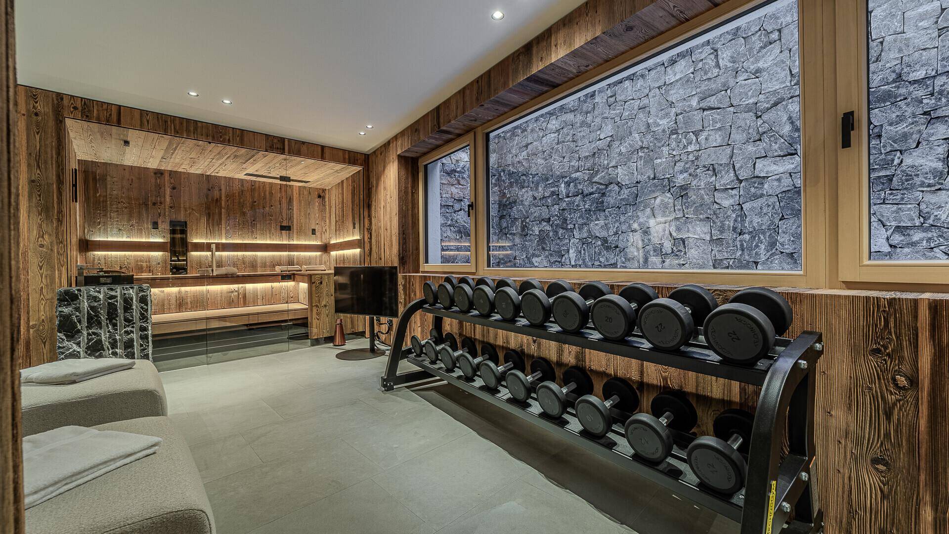 Geräumiger Fitness- und Wellnessbereich mit Holzsauna, Hantelablage und Blick auf eine Steinwand.
