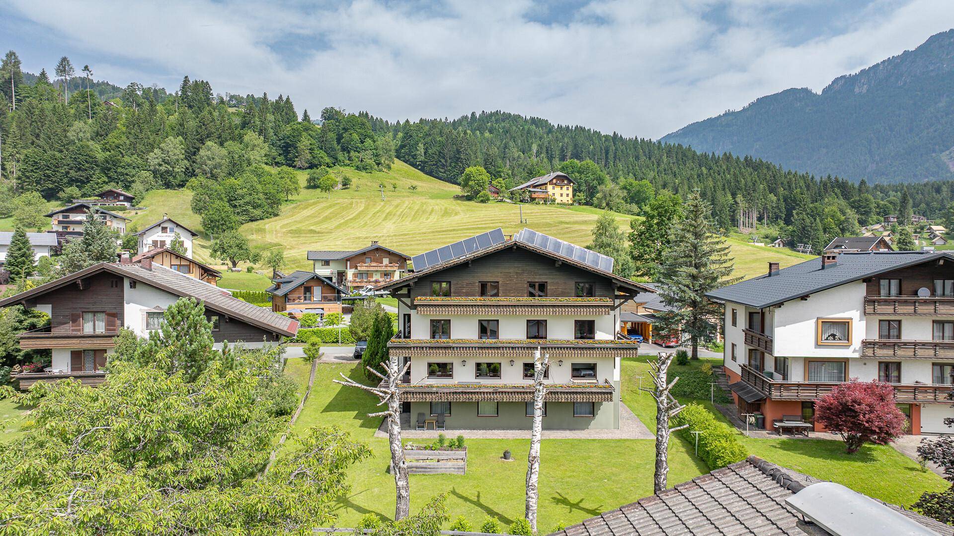 Luftaufnahme eines Gebäudes mit Solarpaneelen und umliegenden Häusern in einer malerischen Bergregion.