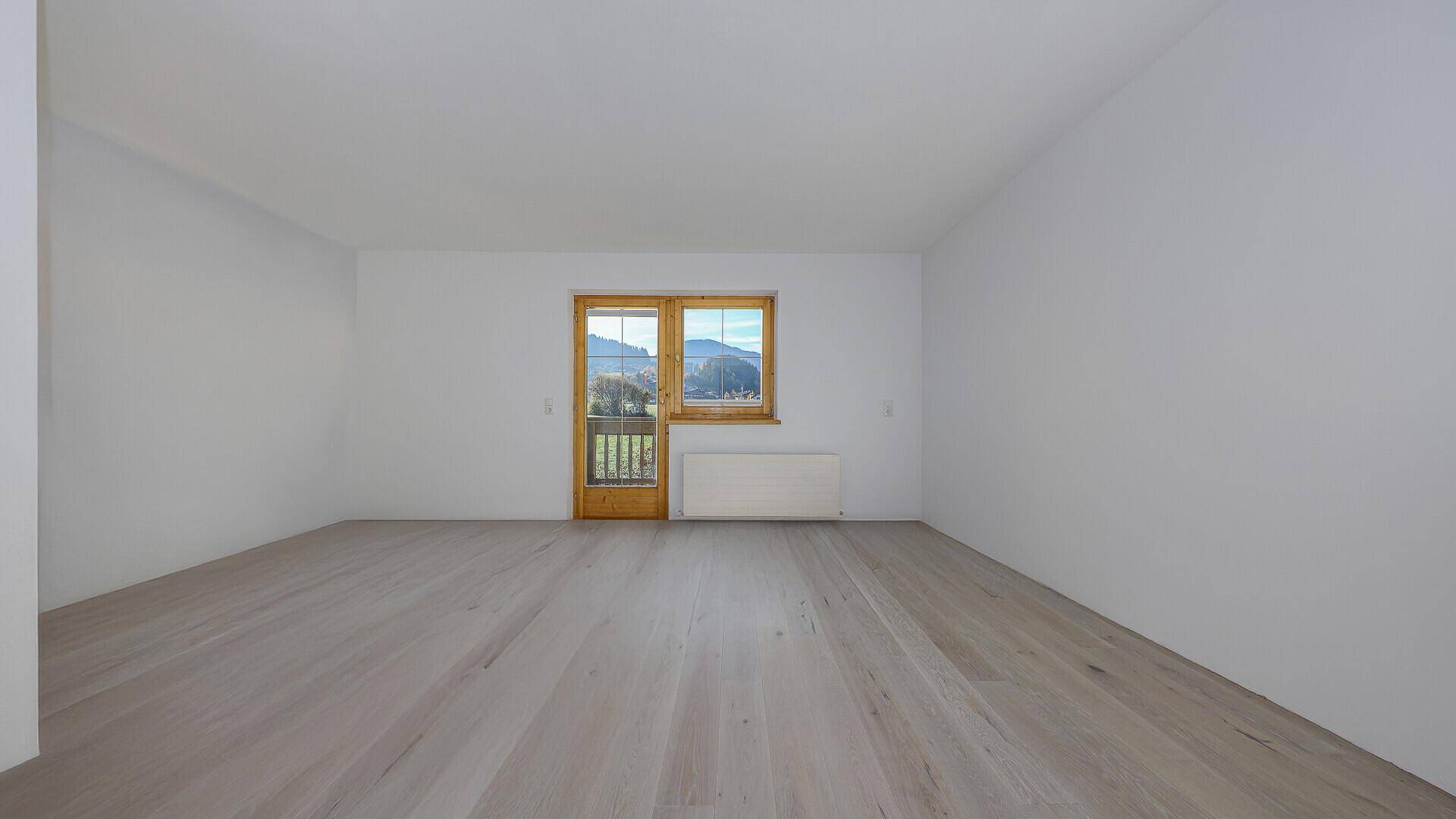 Helles, leeres Wohnzimmer mit Holzboden und Zugang zu einem Balkon mit Bergblick.