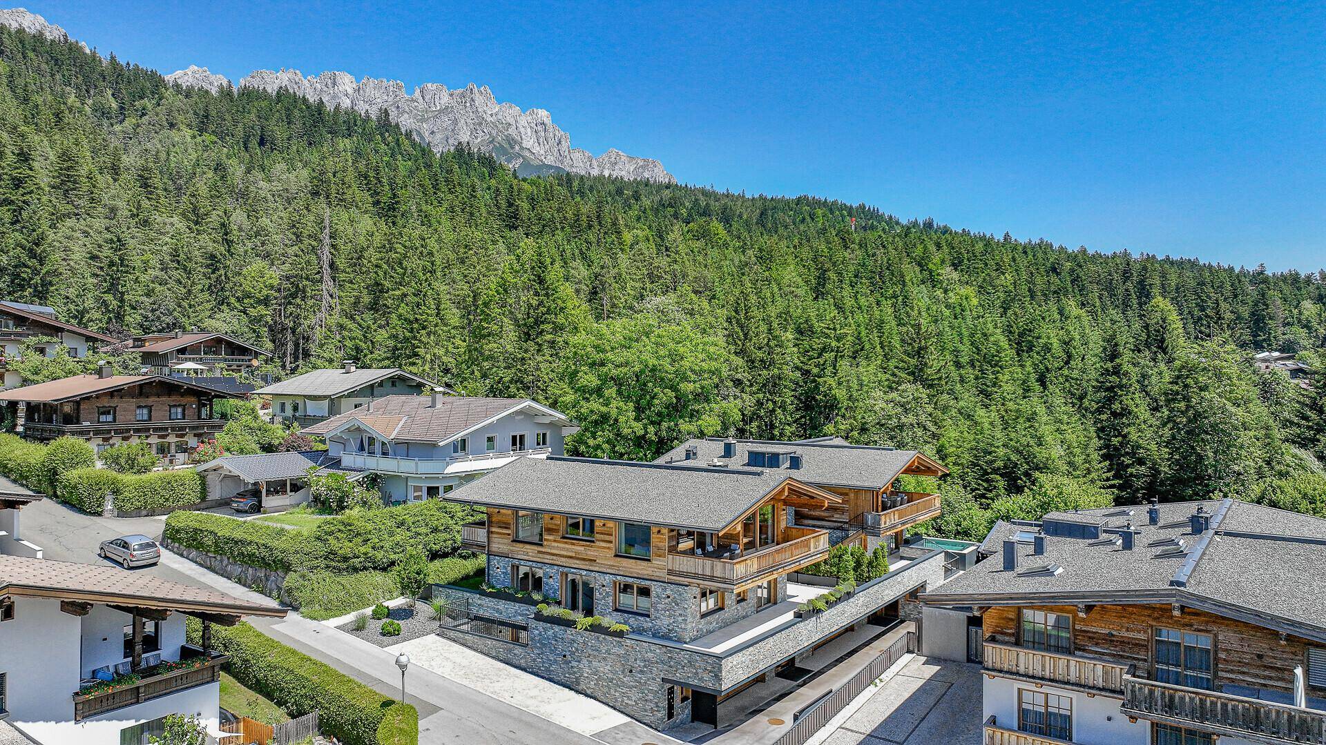 Modernes Chalet in alpiner Umgebung mit Blick auf bewaldete Berge und blauen Himmel.