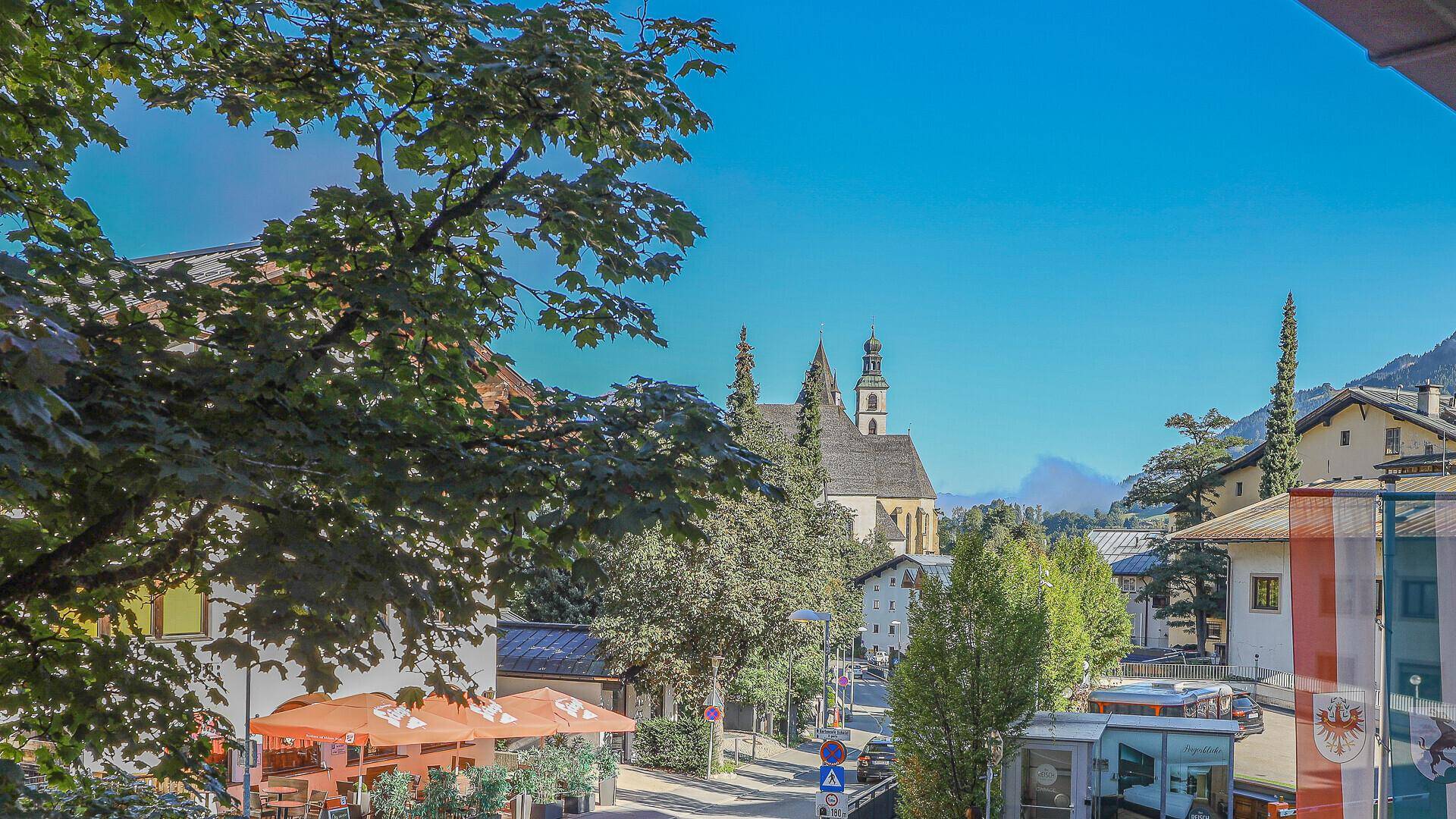 Blick auf eine belebte Straße mit Geschäften, Bäumen und einer Kirche im Hintergrund.