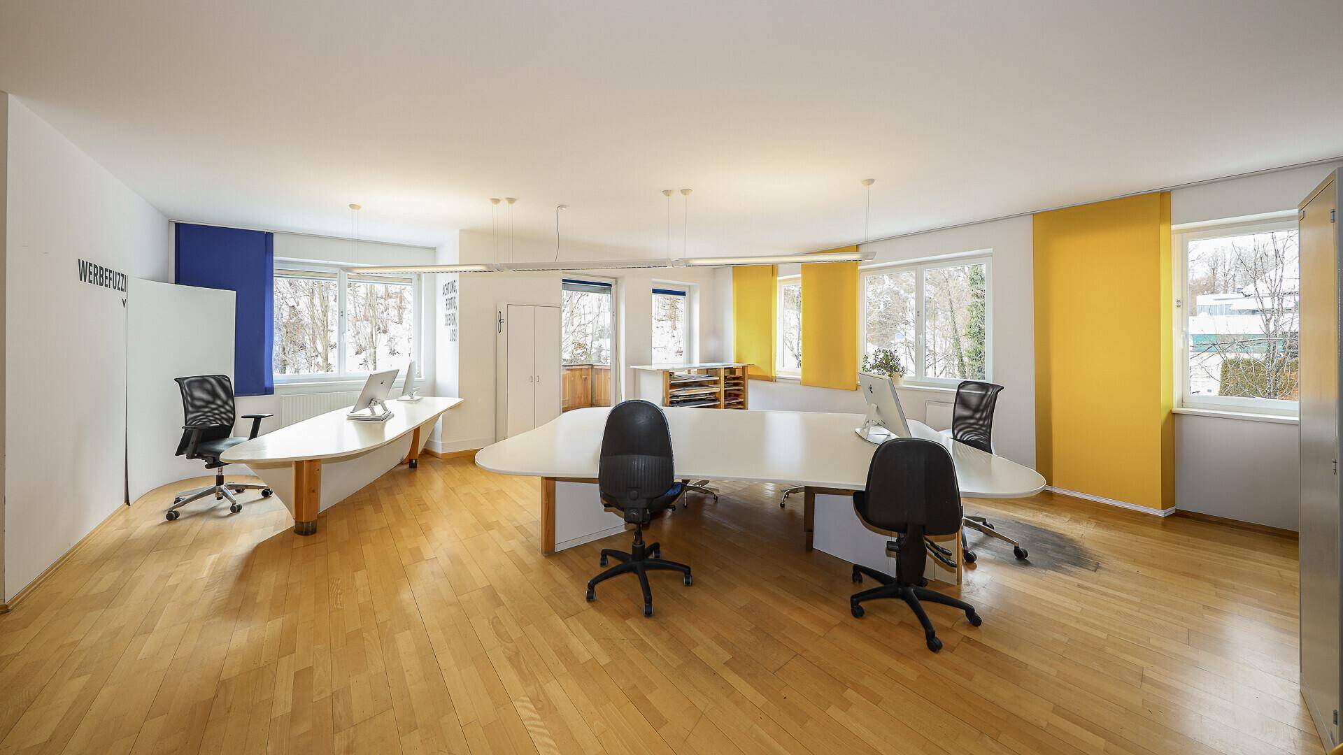 Geräumiges Büro mit Holzboden, gelben und blauen Wänden sowie mehreren Schreibtischen und Bürostühlen.