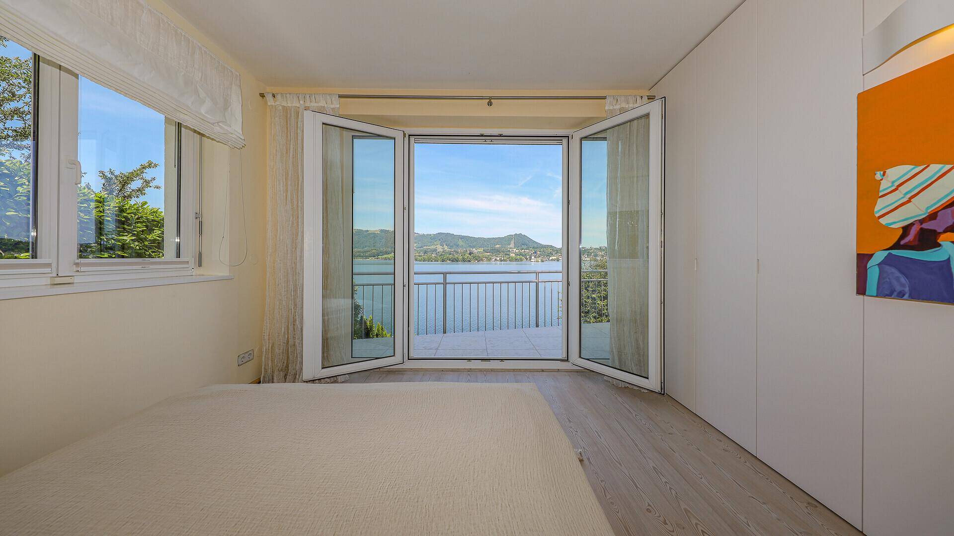 Schlafzimmer mit großen Fenstern, Holzboden und direktem Zugang zu einem Balkon mit Seeblick.