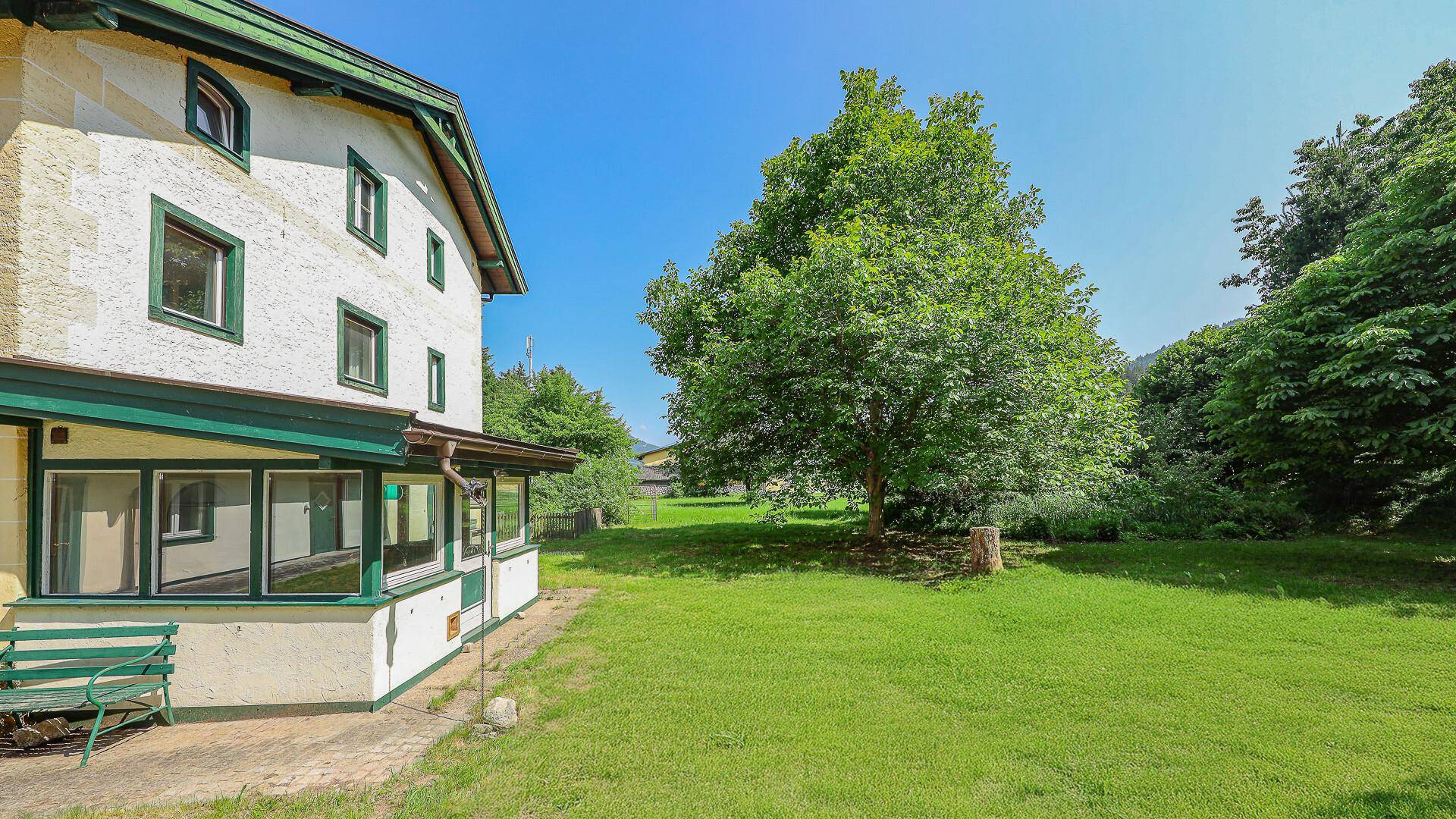 Seitenansicht des Hauses mit großem Rasenbereich und altem Baumbestand unter strahlendem Himmel.