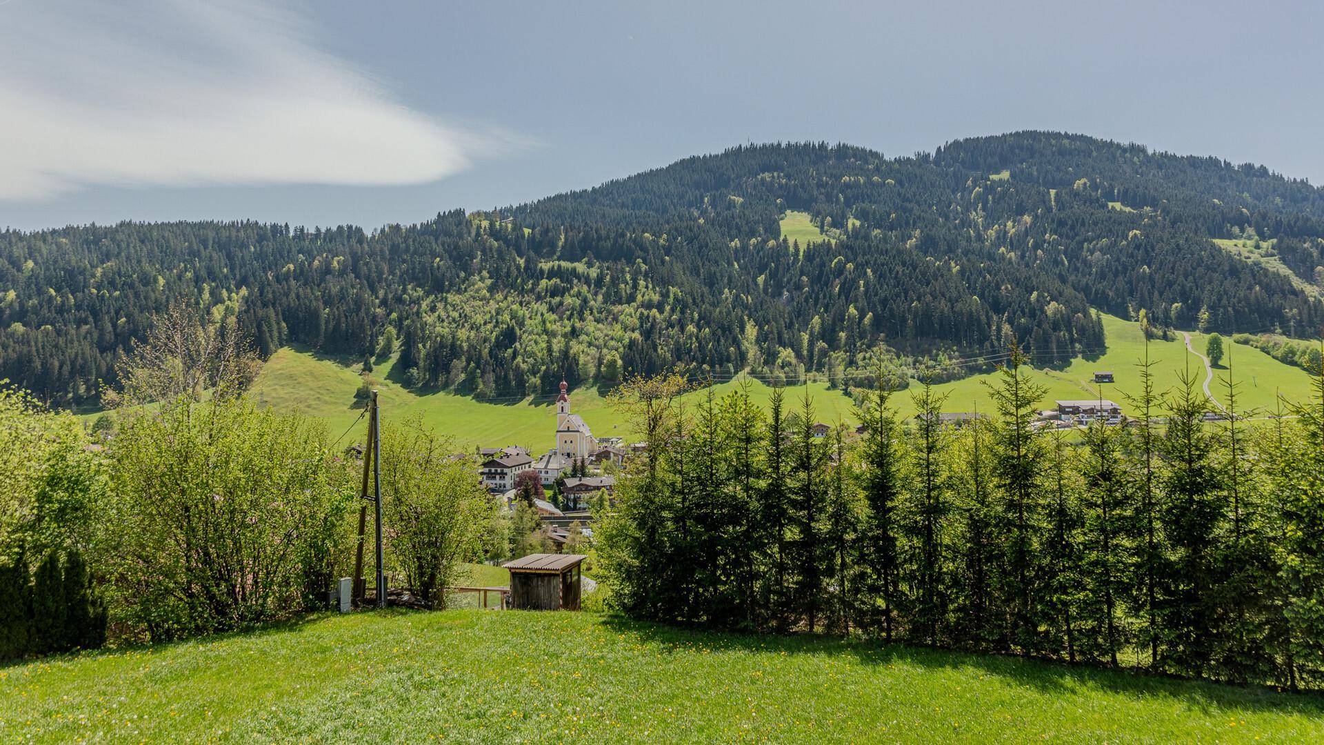 Panoramablick über grüne Wiesen auf ein malerisches Dorf mit Kirchturm und bewaldeten Bergen im Hintergrund.