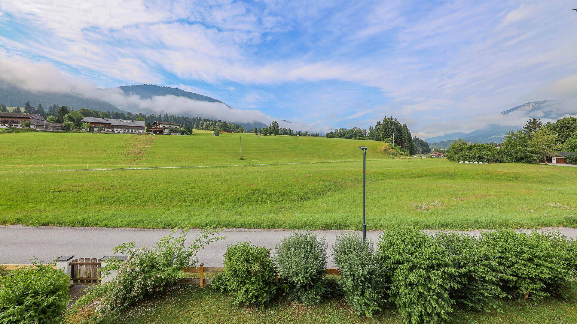 Weitläufiger Blick über grüne Wiesen und sanfte Hügel mit Bergen im Hintergrund unter bewölktem Himmel.