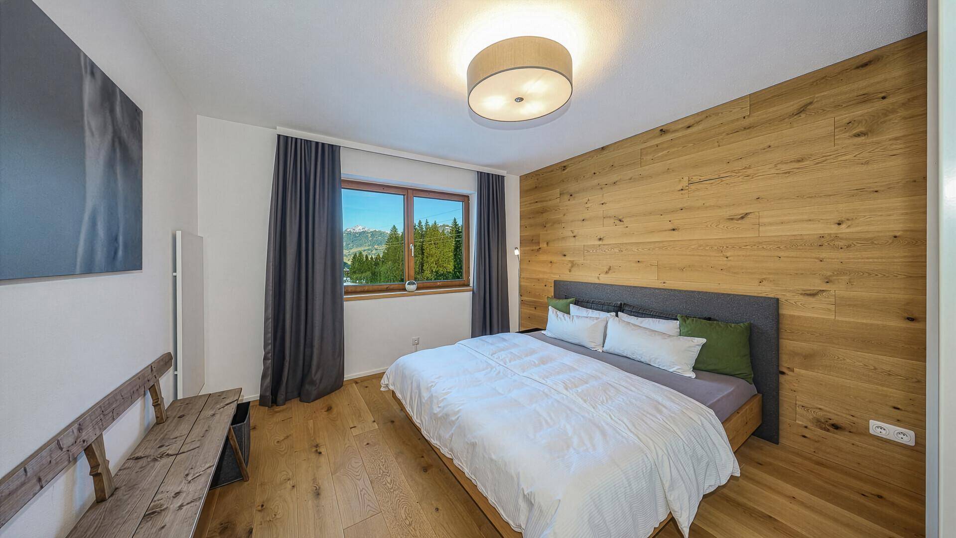 Helles Schlafzimmer mit Holzwand, Doppelbett und Fenster mit Bergblick.
