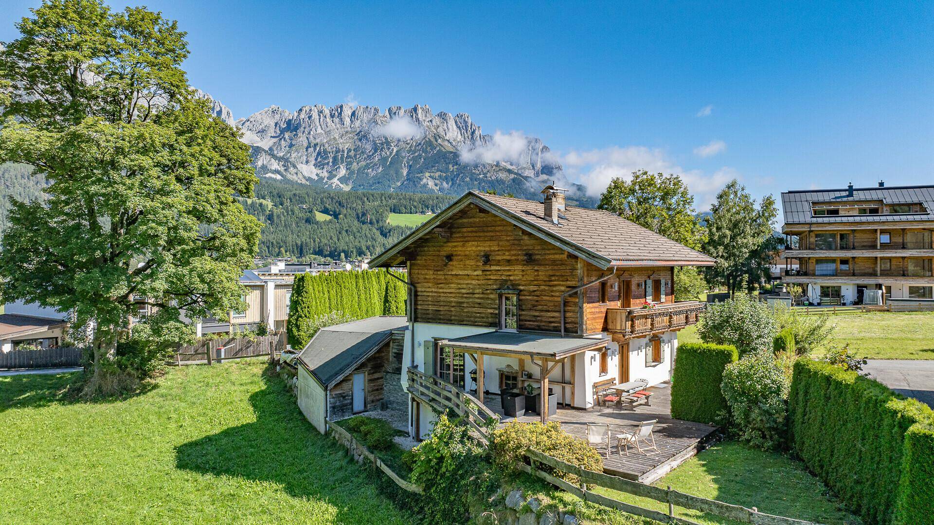 Traditionelles Holzhaus mit Bergblick, umgeben von grünen Wiesen und Bäumen.