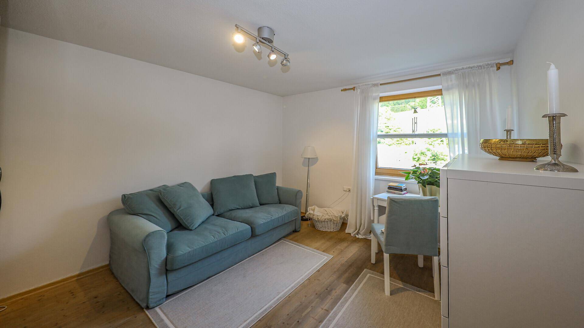 Helles Zimmer mit blauem Sofa, Schreibtisch und Fenster mit Blick ins Grüne.