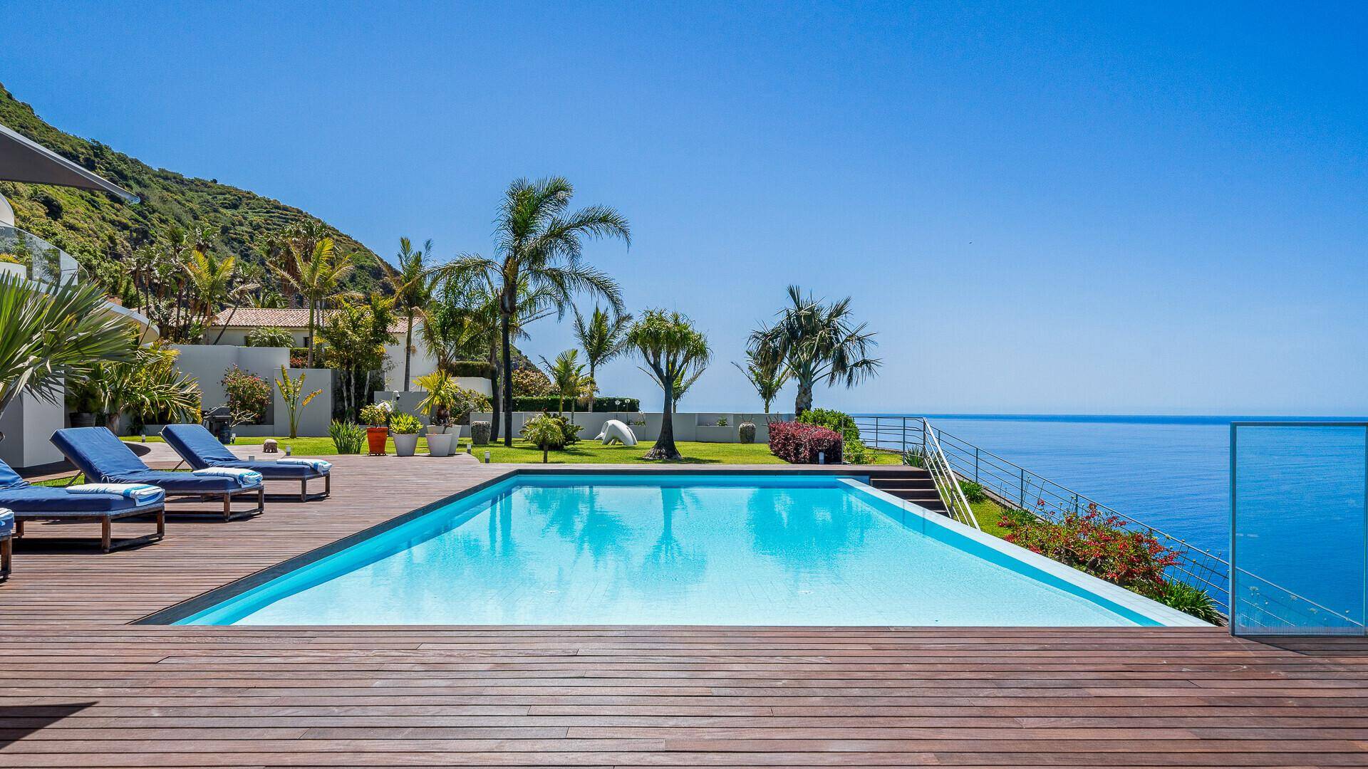 Großer Poolbereich mit Holzdeck und Sonnenliegen, umgeben von Grünflächen mit Blick auf das Meer und den Hang.