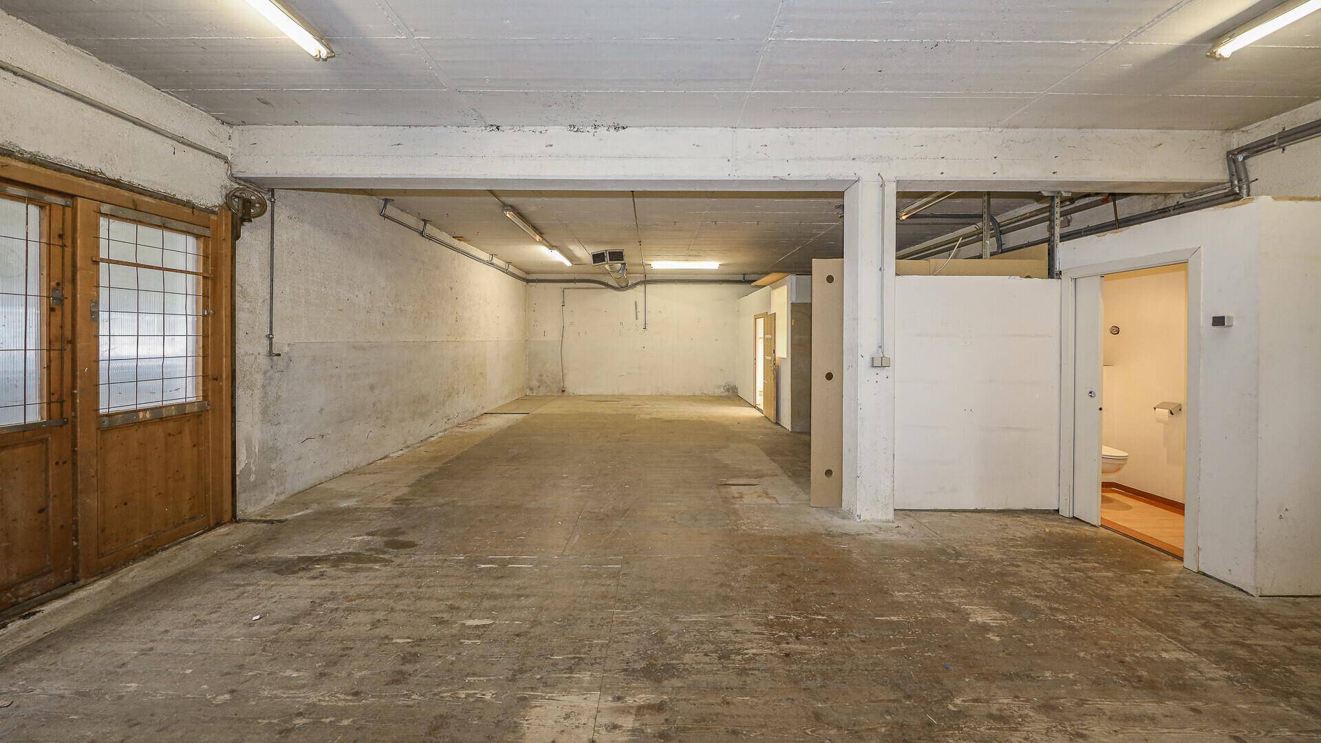 Großer Lagerraum mit Betonboden und Zugang zu einem kleinen Badezimmer, beleuchtet von Deckenleuchten.